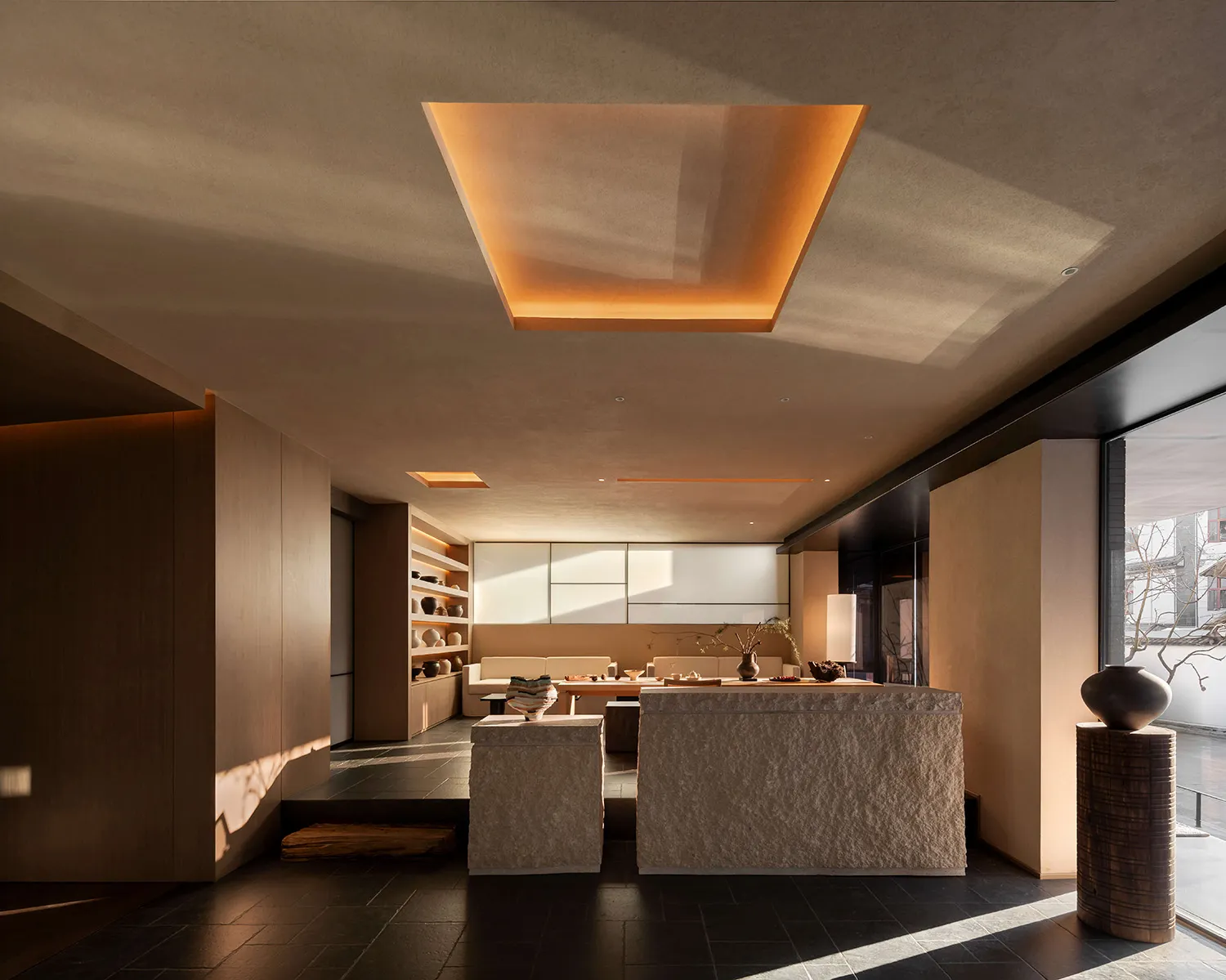 Modern minimalist living room featuring a large travertine central island, sunken seating area, built-in wooden shelving, and a glowing rectangular skylight casting golden light across the dark tiled floor.