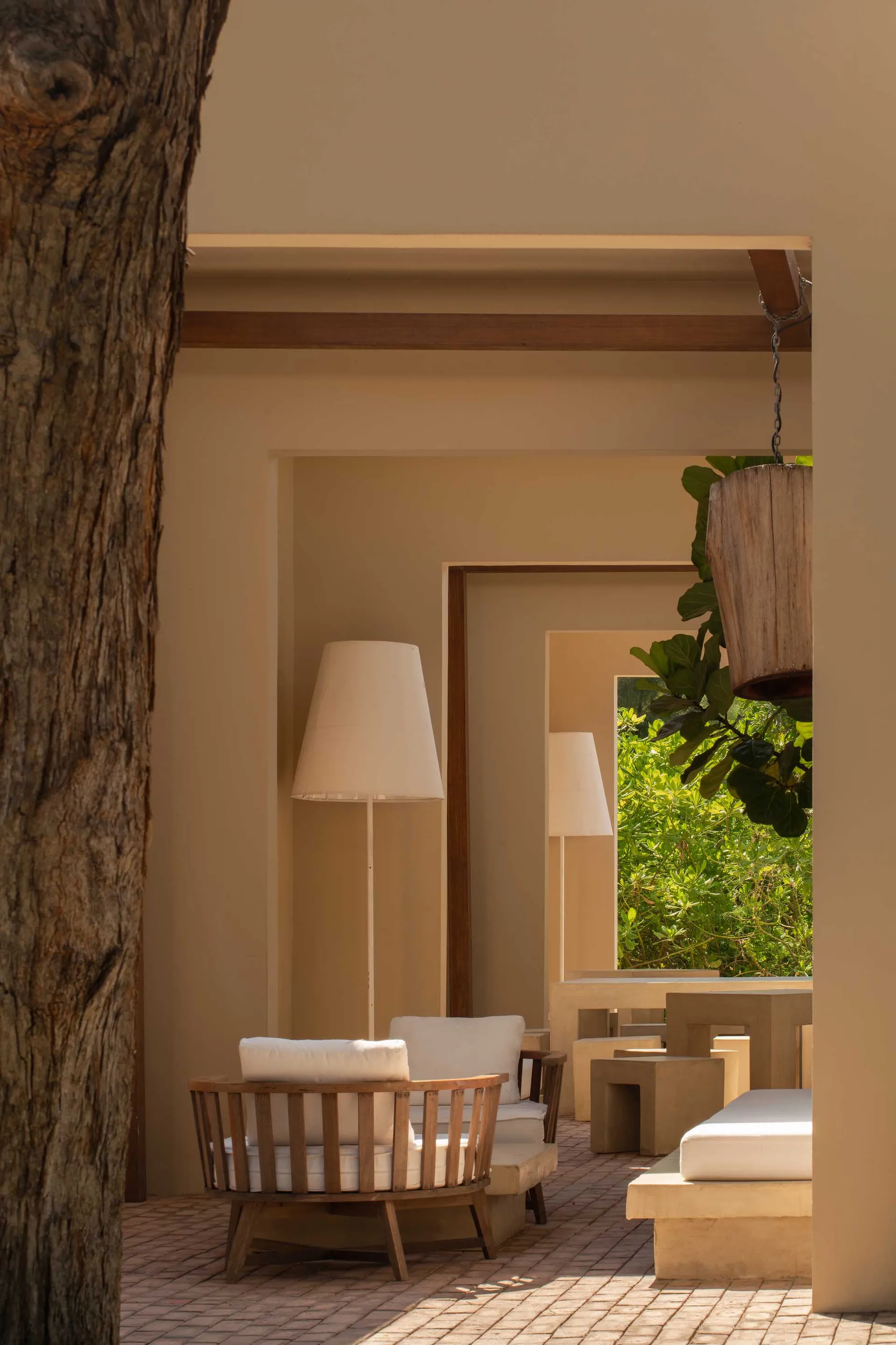 Wooden teak swivel armchair with white cushions placed on a brick-paved courtyard next to concrete outdoor furniture and large floor lamps, framed by a textured tree trunk and lush green foliage in the background.
