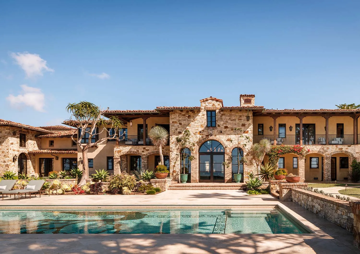 Front view of a luxurious Mediterranean-style stone villa with a rectangular swimming pool in the foreground, arched windows, terracotta roof tiles, and lush tropical landscaping under a clear blue sky.