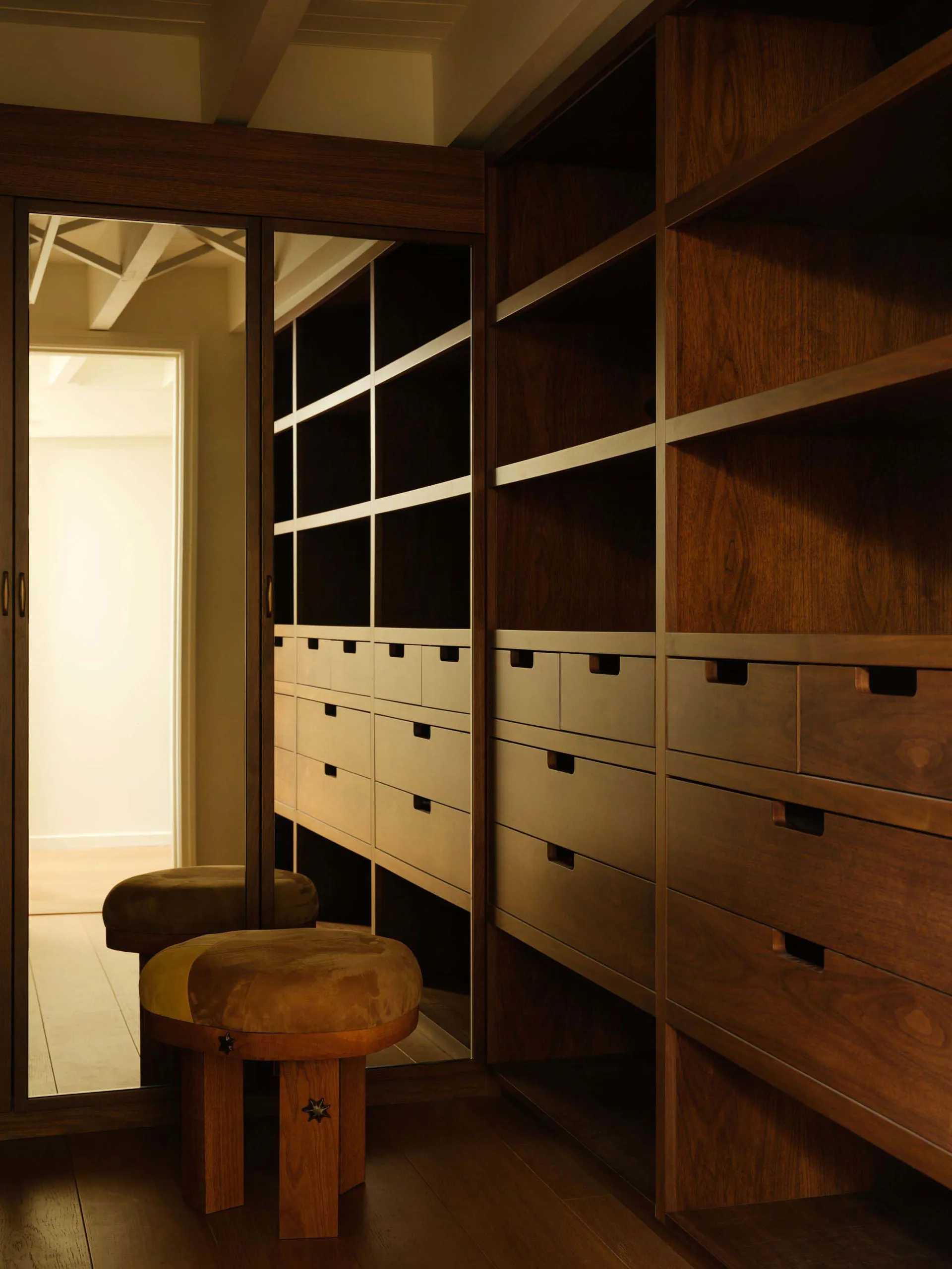 Luxurious walk-in closet featuring rich walnut wood cabinetry, white drawer fronts, full-length mirror doors, and two round velvet ottomans with wooden bases and star-shaped metal details.