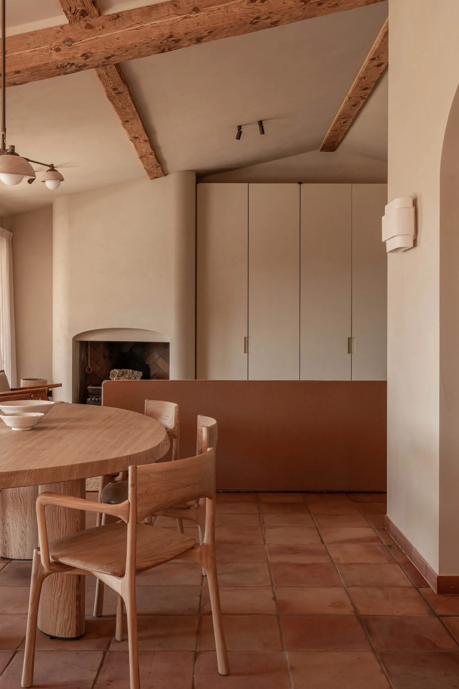 Warm minimalist dining room with round oak table, wooden chairs, terracotta floor tiles, and a fireplace in the background under exposed wooden ceiling beams.