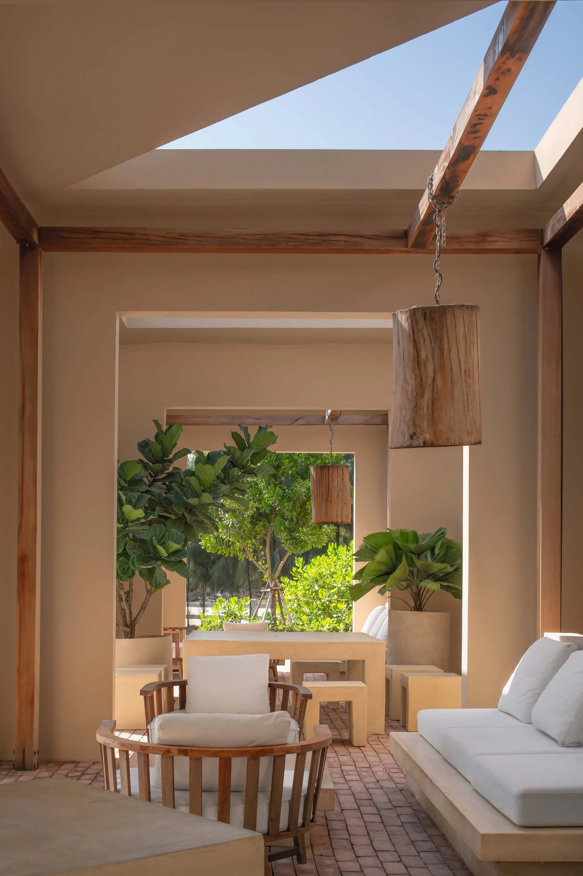 Open-air courtyard patio featuring wooden ceiling beams, a cylindrical wooden pendant lamp, teak armchair with white cushions, built-in stone sofa, and lush potted plants under a bright blue sky.