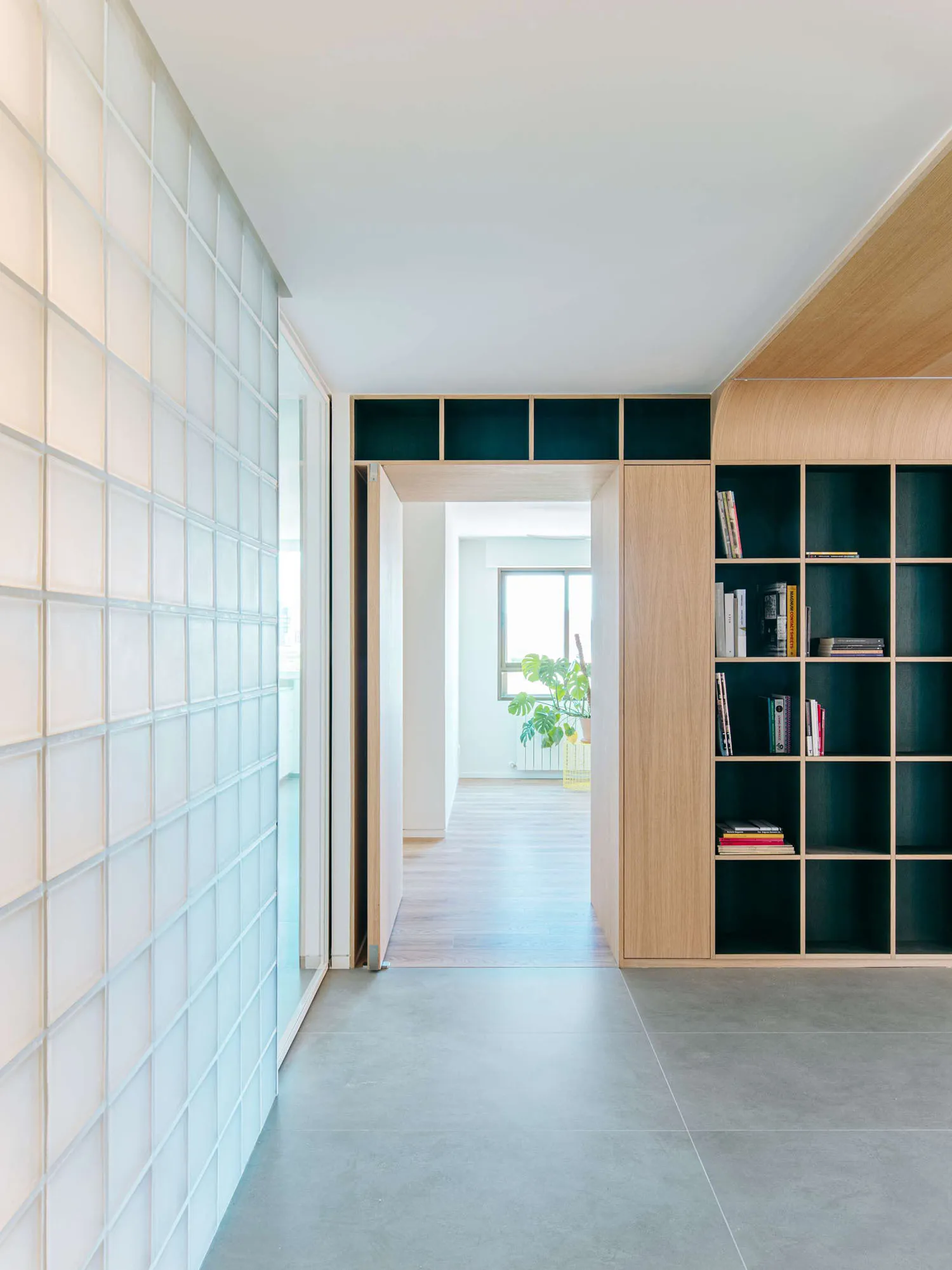 Modern minimalist hallway with light oak built-in bookshelf, white glass block wall, concrete floor, and view into a bright living area with plants.