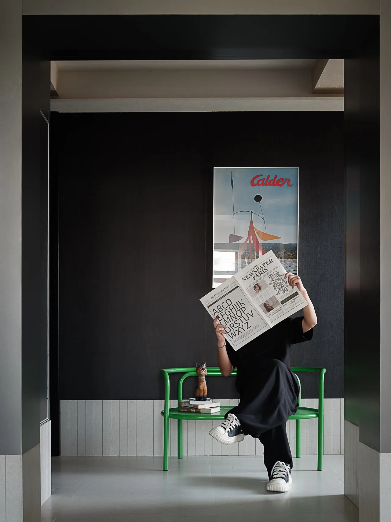 Person reading a newspaper while sitting on a bright green chair in a modern dark entryway with a large Alexander Calder poster on the wall.