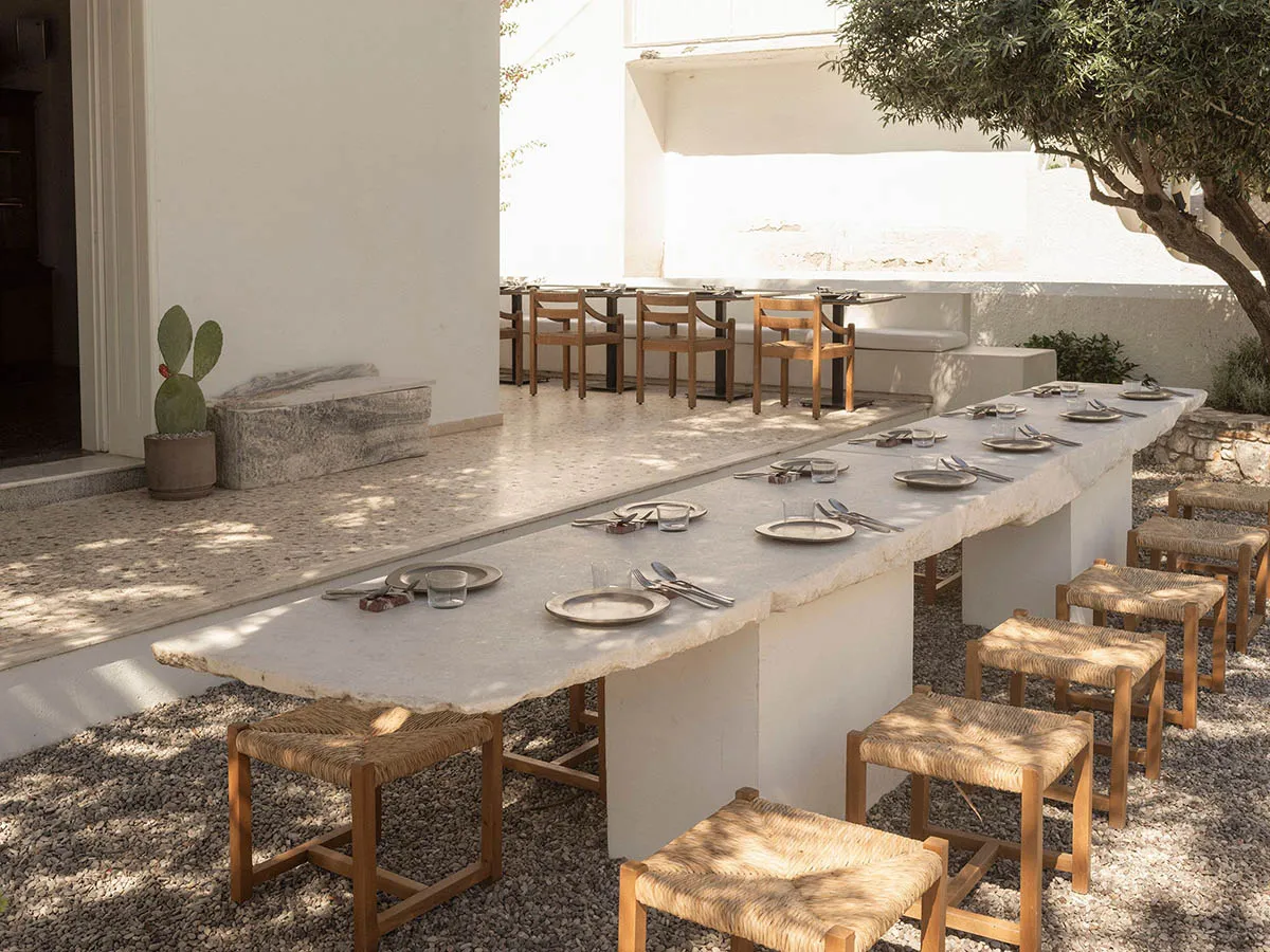 Long outdoor dining table made from natural travertine stone with woven rush stools arranged on gravel in a sunny white-walled Mediterranean courtyard with a prickly pear cactus.
