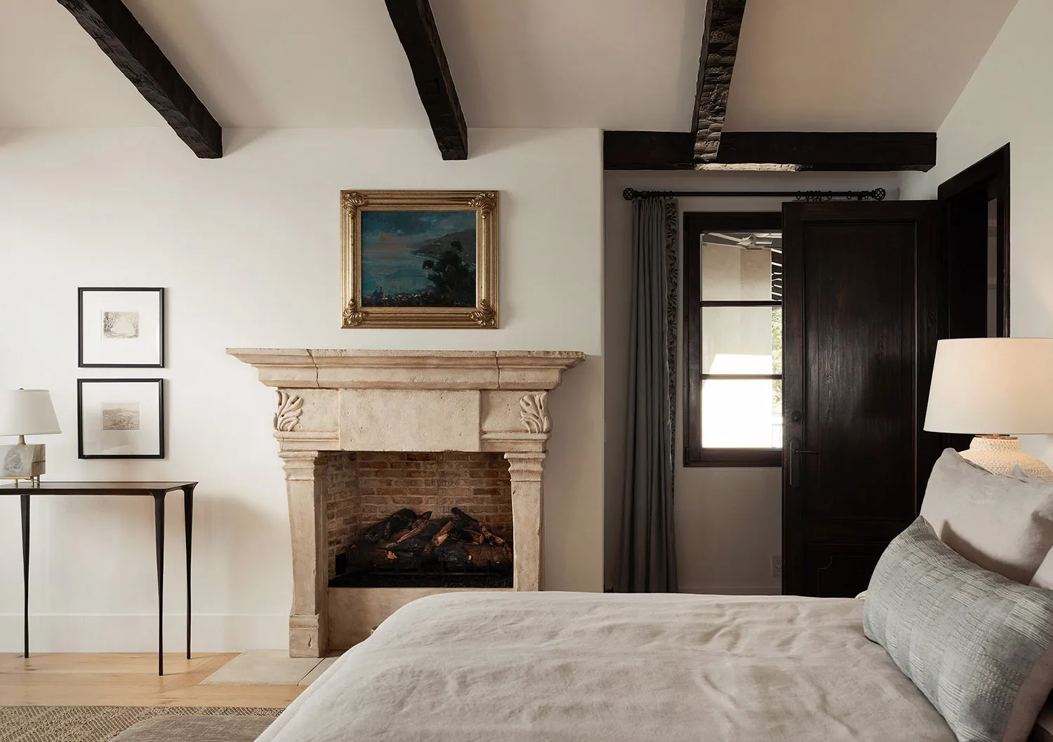 Cozy bedroom interior with a large bed with neutral linen bedding, an antique carved limestone fireplace with brick interior, dark wooden ceiling beams, and framed artwork on white walls.