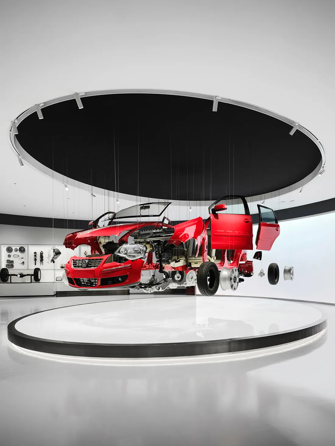 Red Volkswagen car cutaway suspended by wires in a modern white circular exhibition hall with dramatic black circular ceiling feature.