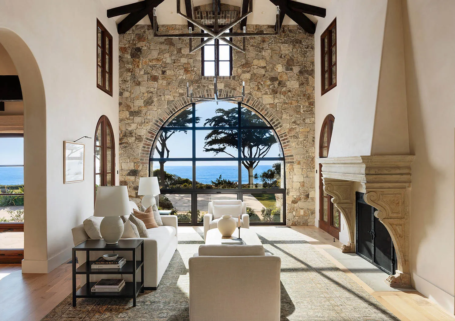 Spacious coastal living room featuring a large stone fireplace, white sofas, high beamed ceilings, and a dramatic arched picture window overlooking the ocean and trees.