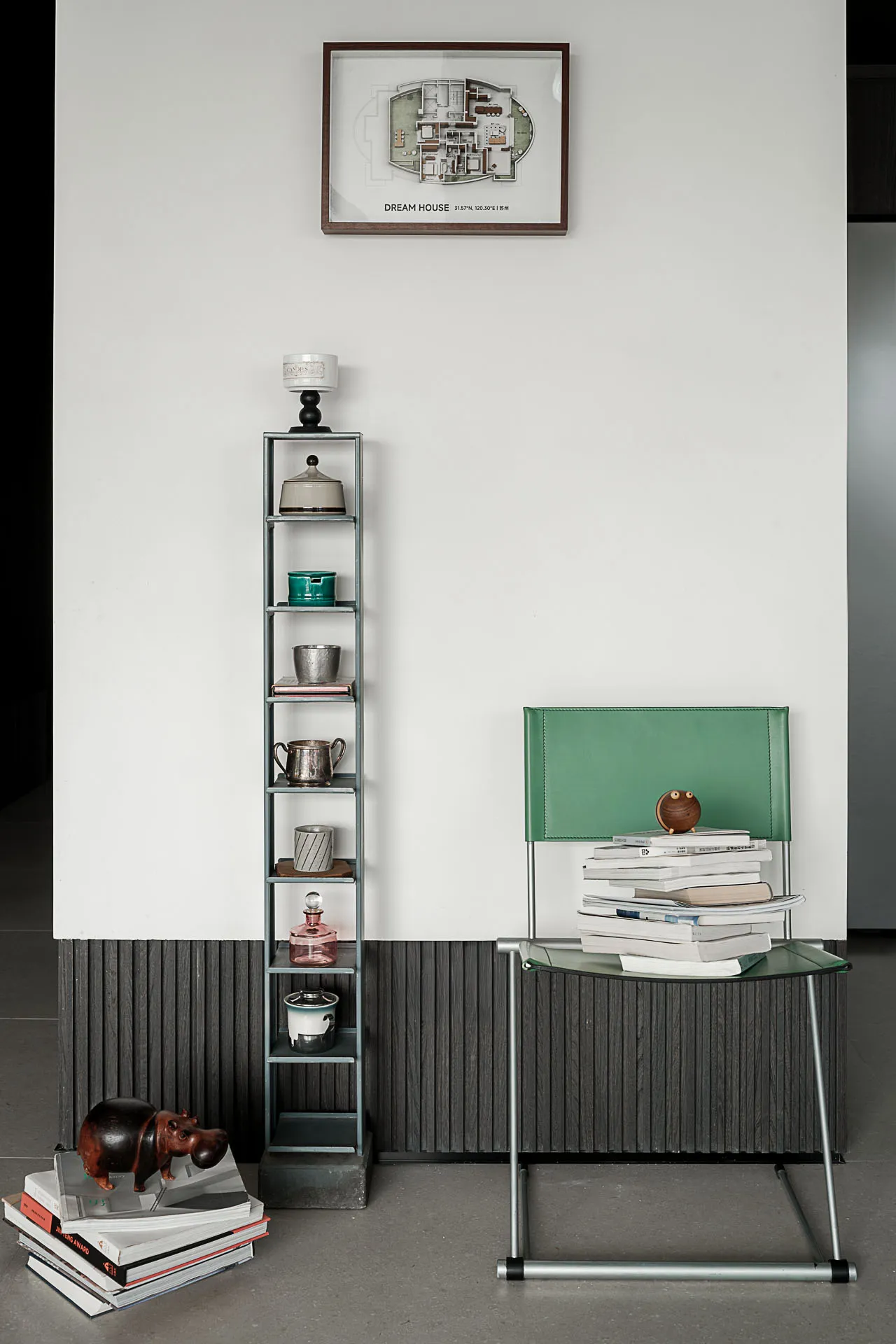 Modern minimalist entryway featuring a tall grey metal shelving unit displaying vintage objects, a bright green leather chair with stacked books, and a framed architectural floor plan labeled 'DREAM HOUSE' on a white wall with dark wood wainscoting.