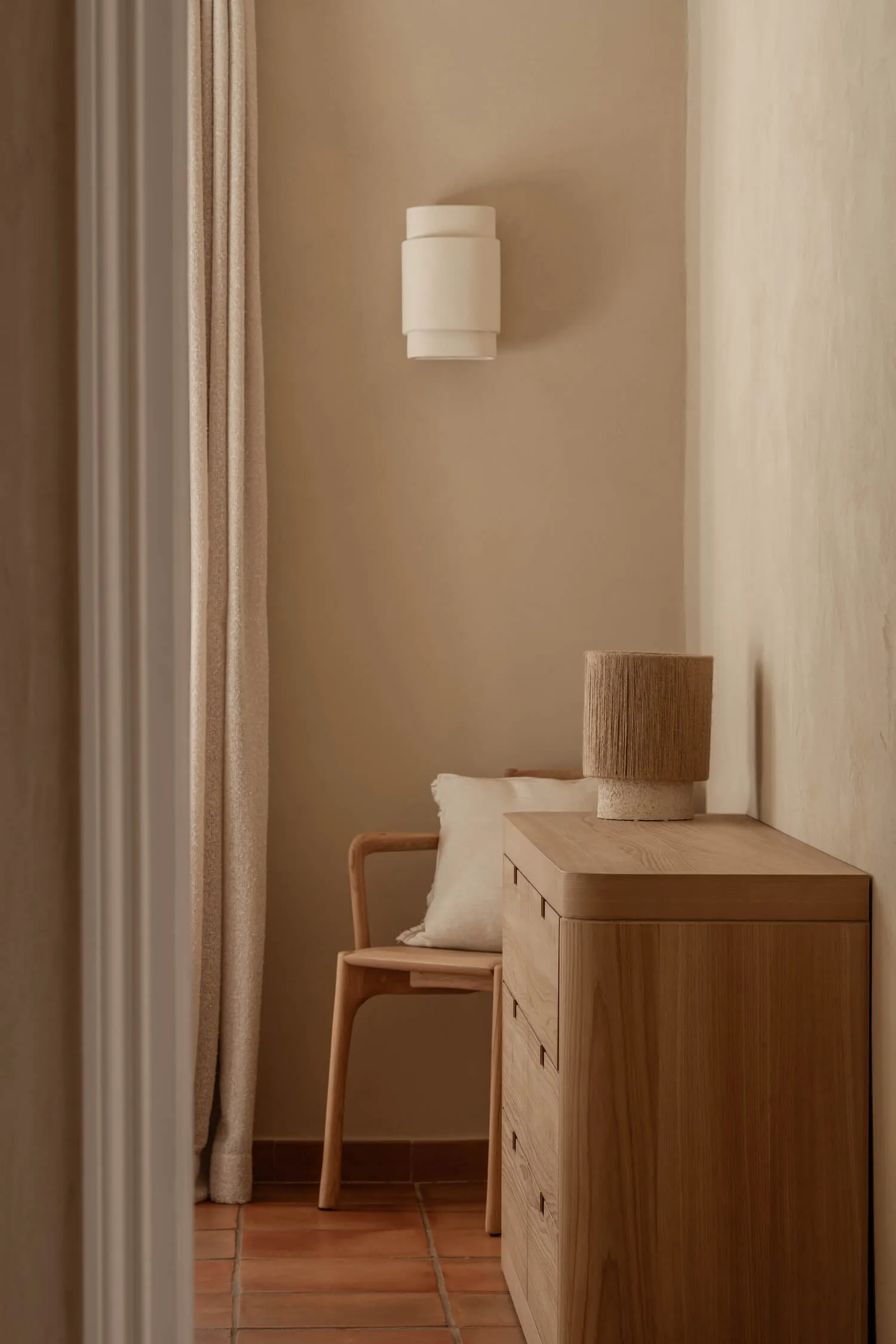 Minimalist entryway featuring light oak dresser, wooden armchair with linen cushion, cylindrical wall sconce, and terracotta tile floor in warm neutral tones.