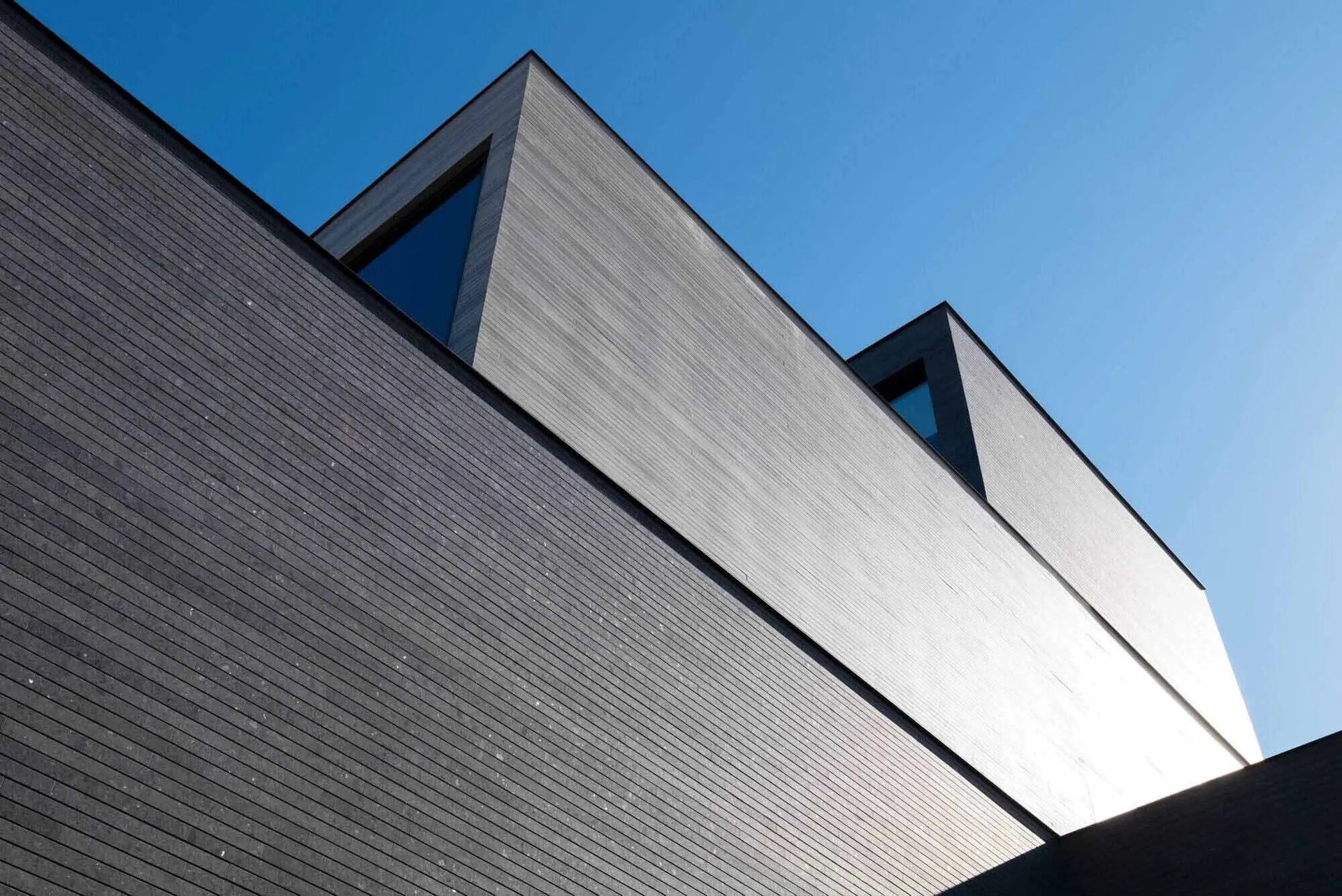 Low-angle view of a contemporary building with dark horizontal wood cladding and distinctive triangular windows set against a clear blue sky.