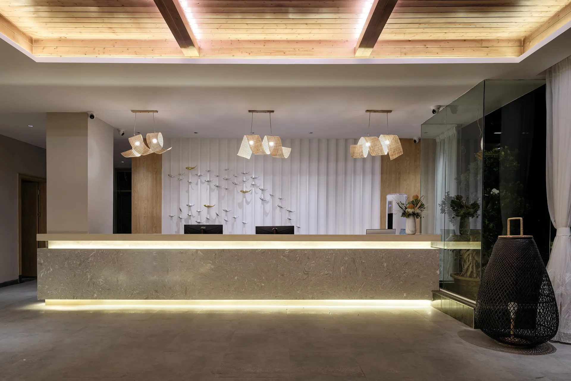 Modern hotel reception desk made of travertine stone with under-lighting, woven rattan pendant lamps, white pleated wall, metallic bird wall art, and wooden ceiling.