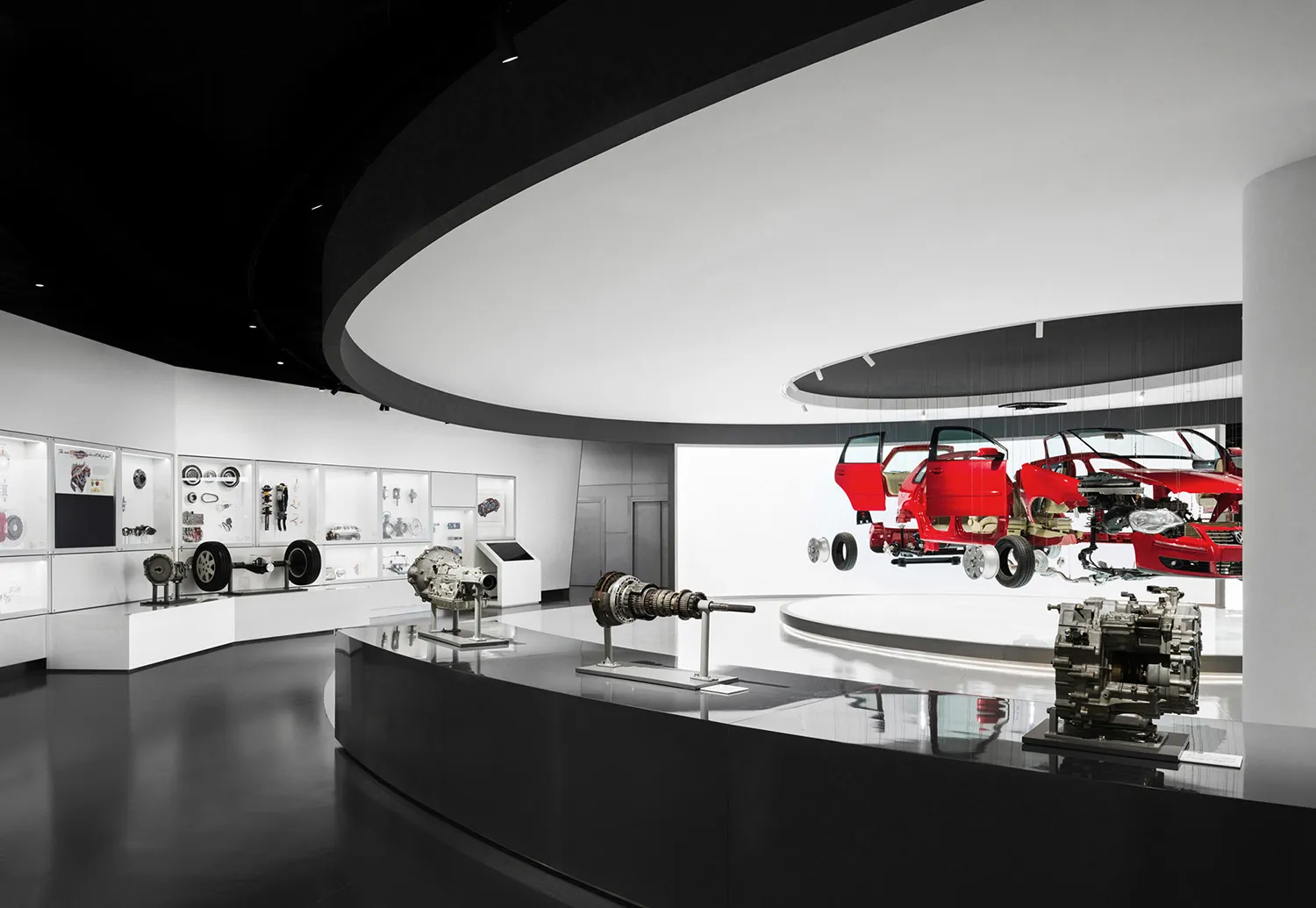 Interior view of a modern automotive museum with suspended red cutaway car sections, large mechanical components on glossy black pedestals, and illuminated display cases along curved white walls.