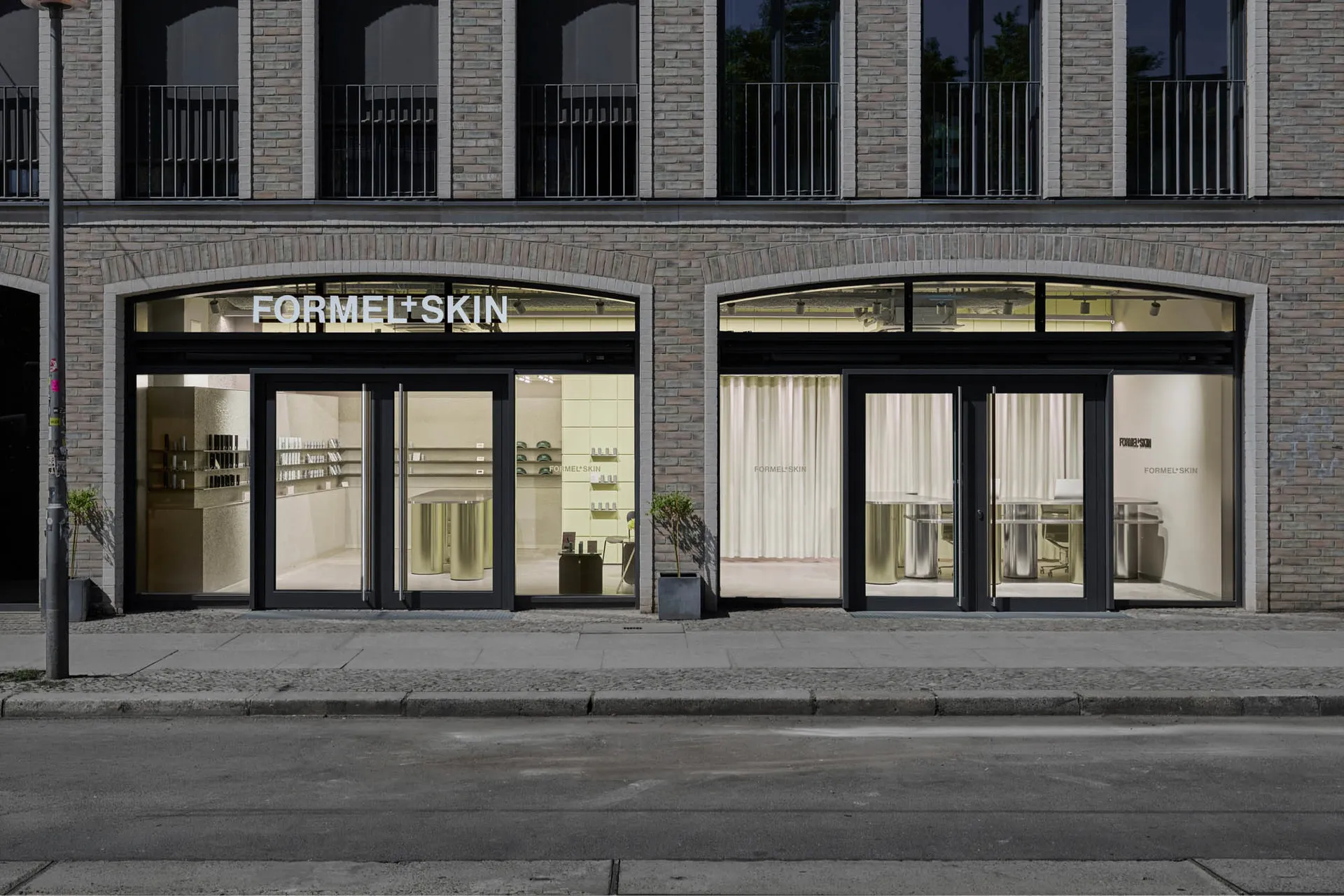 柏林 Formel Skin 美容診所店面街景,兩個拱形玻璃入口搭配黑色框架、黃銅圓柱展示桌、淡黃色內牆,以及人行道上的盆栽樹木。