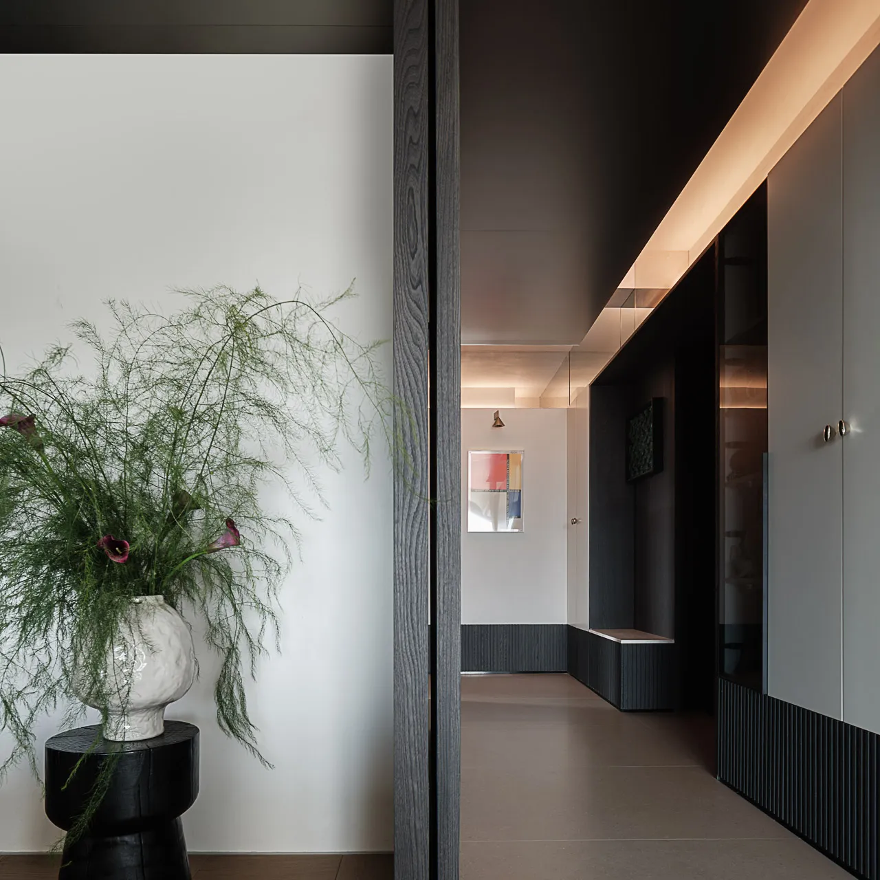 Modern minimalist hallway featuring dark wood paneling, built-in storage, warm linear LED lighting, and a large floral arrangement on a black side table.