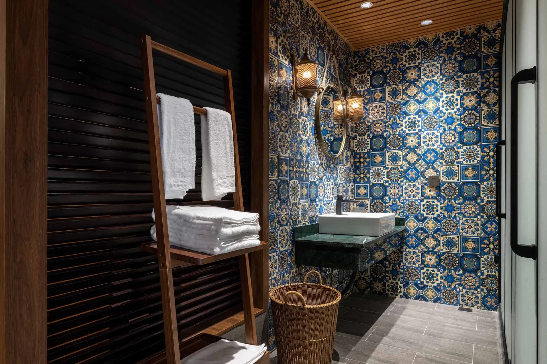 Compact powder room featuring vibrant blue and gold Moroccan patterned tiles, white vessel sink on green marble countertop, round mirror with wall sconces, wooden ladder towel rack with white towels, and woven basket on dark grey floor tiles.