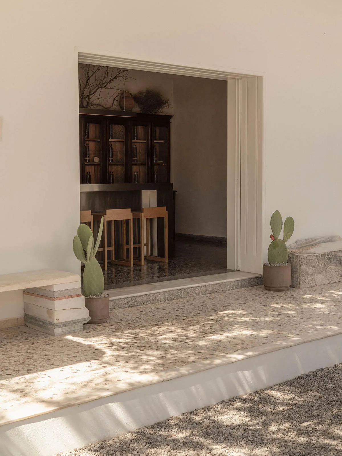 View from a sunlit terrazzo outdoor terrace with potted cacti and stacked stone bench, looking through an open doorway into a dark wood bar area with wooden stools.