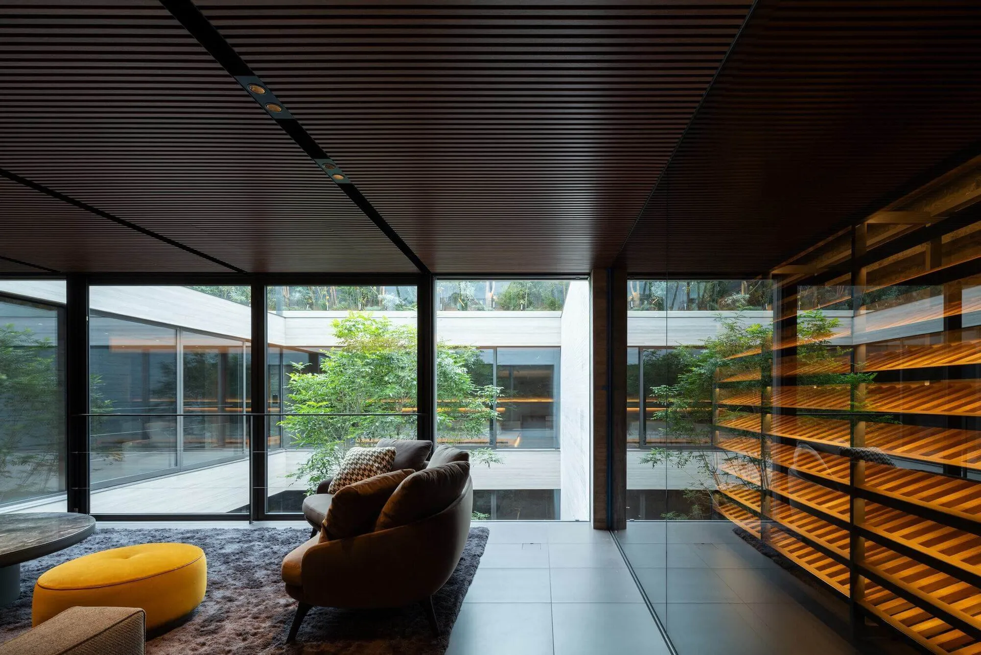 Modern living room featuring a dark wood slatted ceiling, large glass walls overlooking a garden, brown curved sofa, bright yellow ottoman, and illuminated wooden shelving unit.