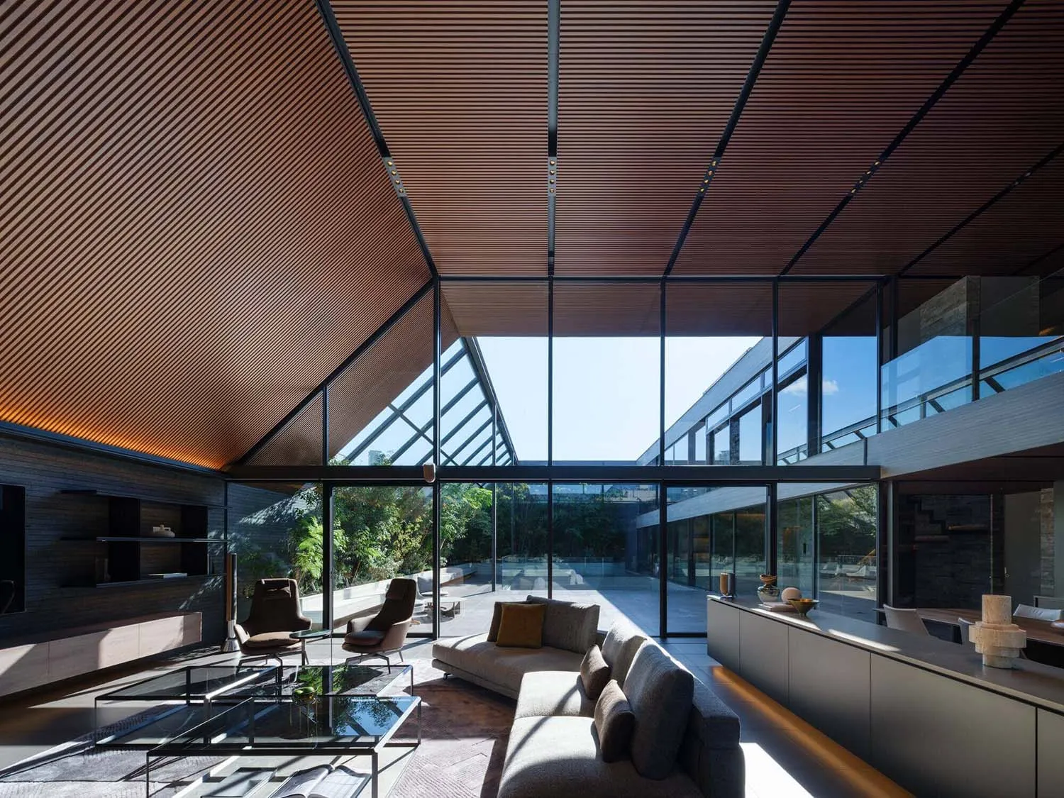 Modern double-height living room featuring a slatted wood ceiling, expansive glass walls overlooking a garden, a grey L-shaped sectional sofa, and minimalist furniture under bright natural light.