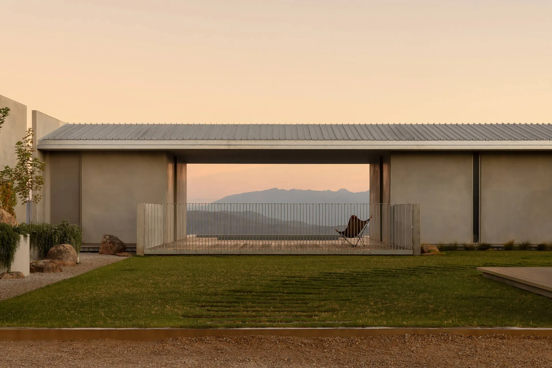 Modern concrete house with open patio, butterfly chair, wooden deck, and distant mountain range at sunset