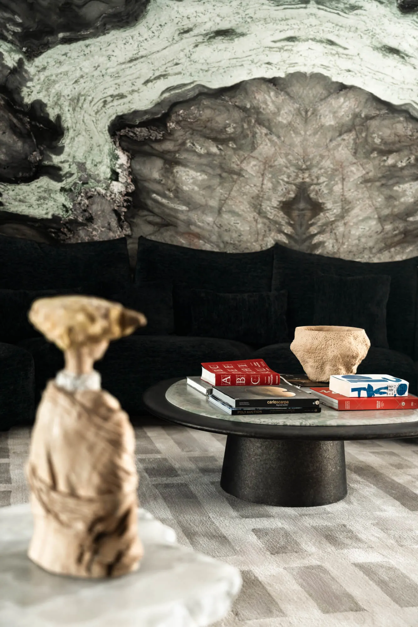 Modern living room featuring a large abstract green and grey marble wall, black velvet sectional sofa with multiple cushions, round dark coffee table with books and stone bowl, and foreground stone sculpture.
