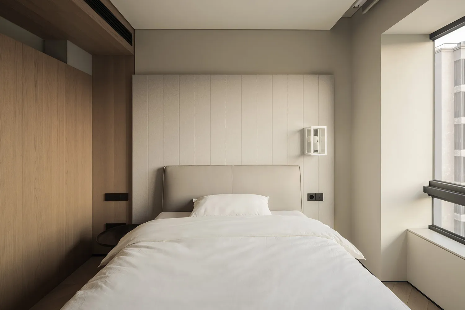 Modern minimalist bedroom with a beige upholstered bed, white linens, vertical wood panel accent wall, and large window with natural light.