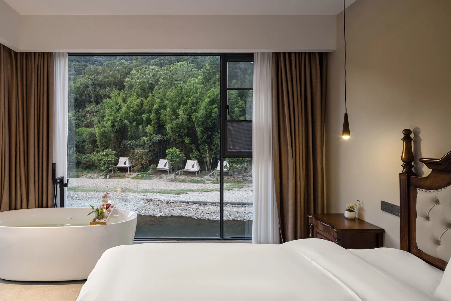 Modern hotel bedroom featuring a large white freestanding bathtub positioned directly in front of a floor-to-ceiling window overlooking a lush green forest and river, with a wooden four-poster bed and dark wood nightstand.