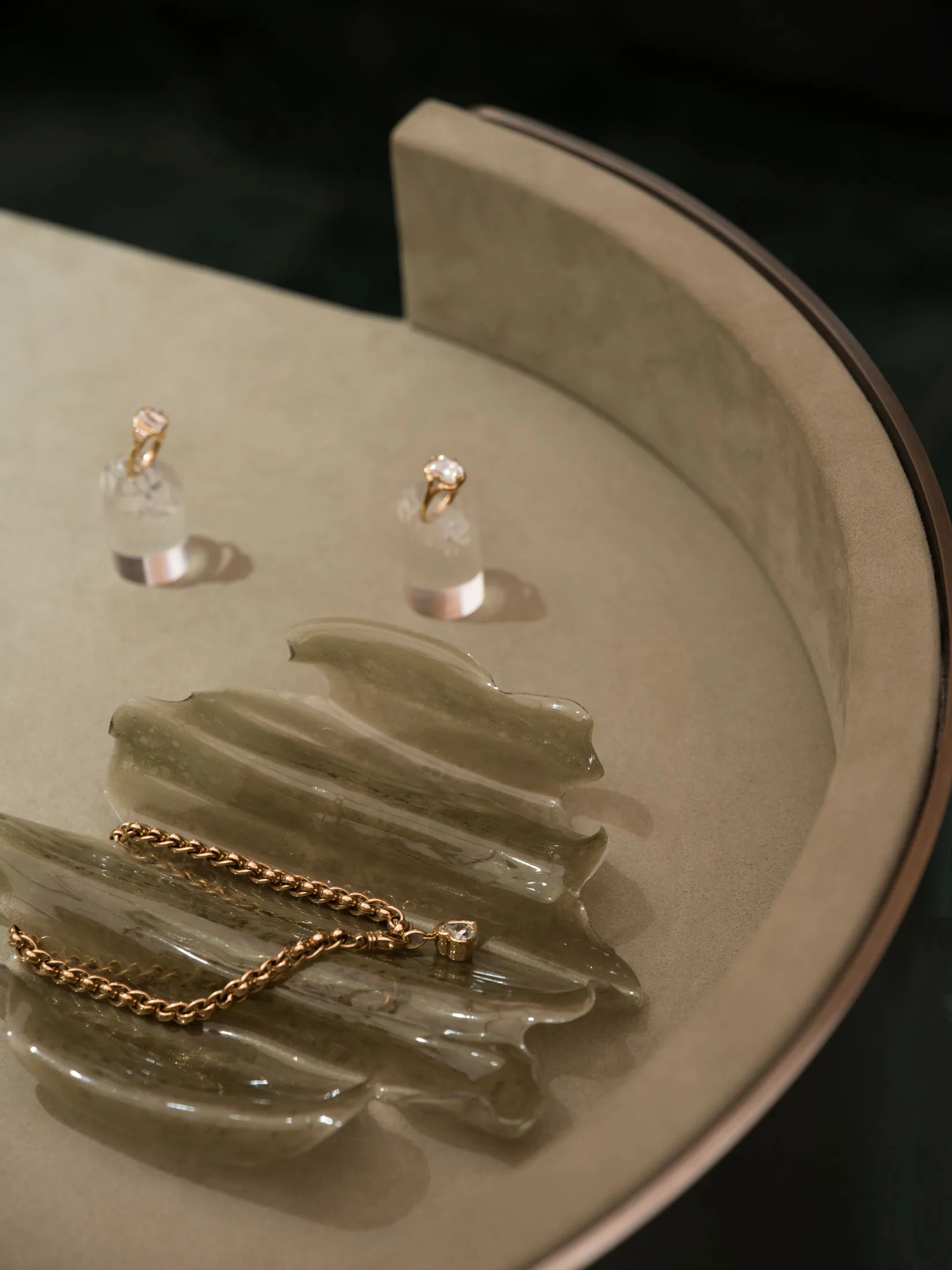 Close-up of a translucent olive-green resin leaf-shaped tray holding a gold rope chain with heart-shaped diamond pendant, accompanied by two diamond solitaire rings displayed on clear acrylic stands, all placed on a curved beige upholstered surface.