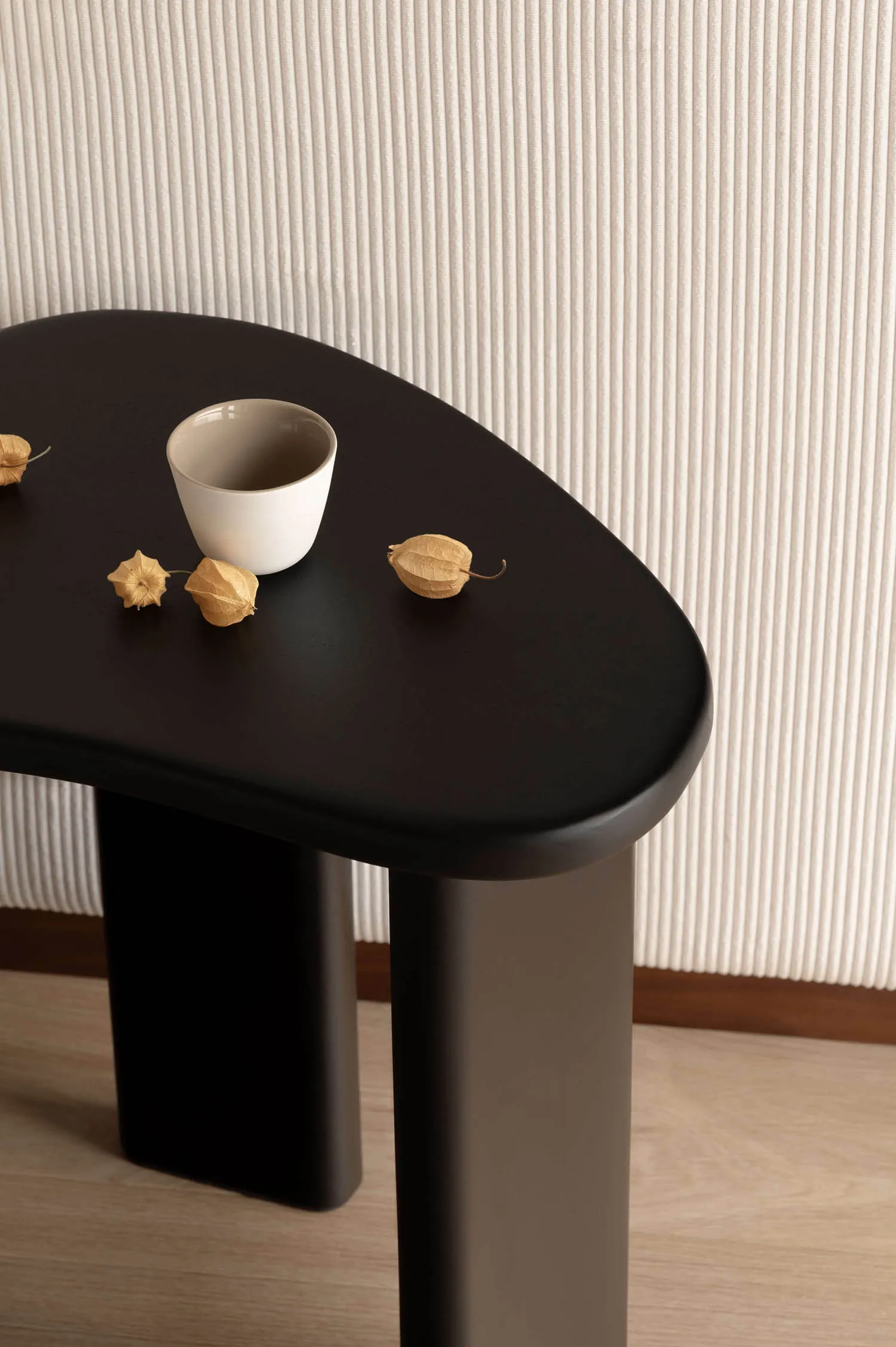 Close-up of a matte black console table with rounded edges, holding a white ceramic bowl and three dried physalis fruits against a vertically ribbed beige wall.