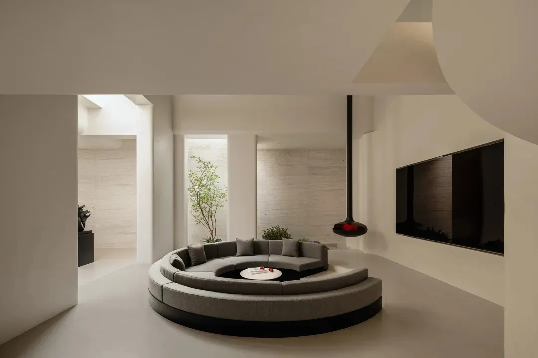 Modern minimalist living room featuring a large grey curved modular sofa on a circular platform, limestone walls, high ceilings with skylights, and a black pendant fireplace.