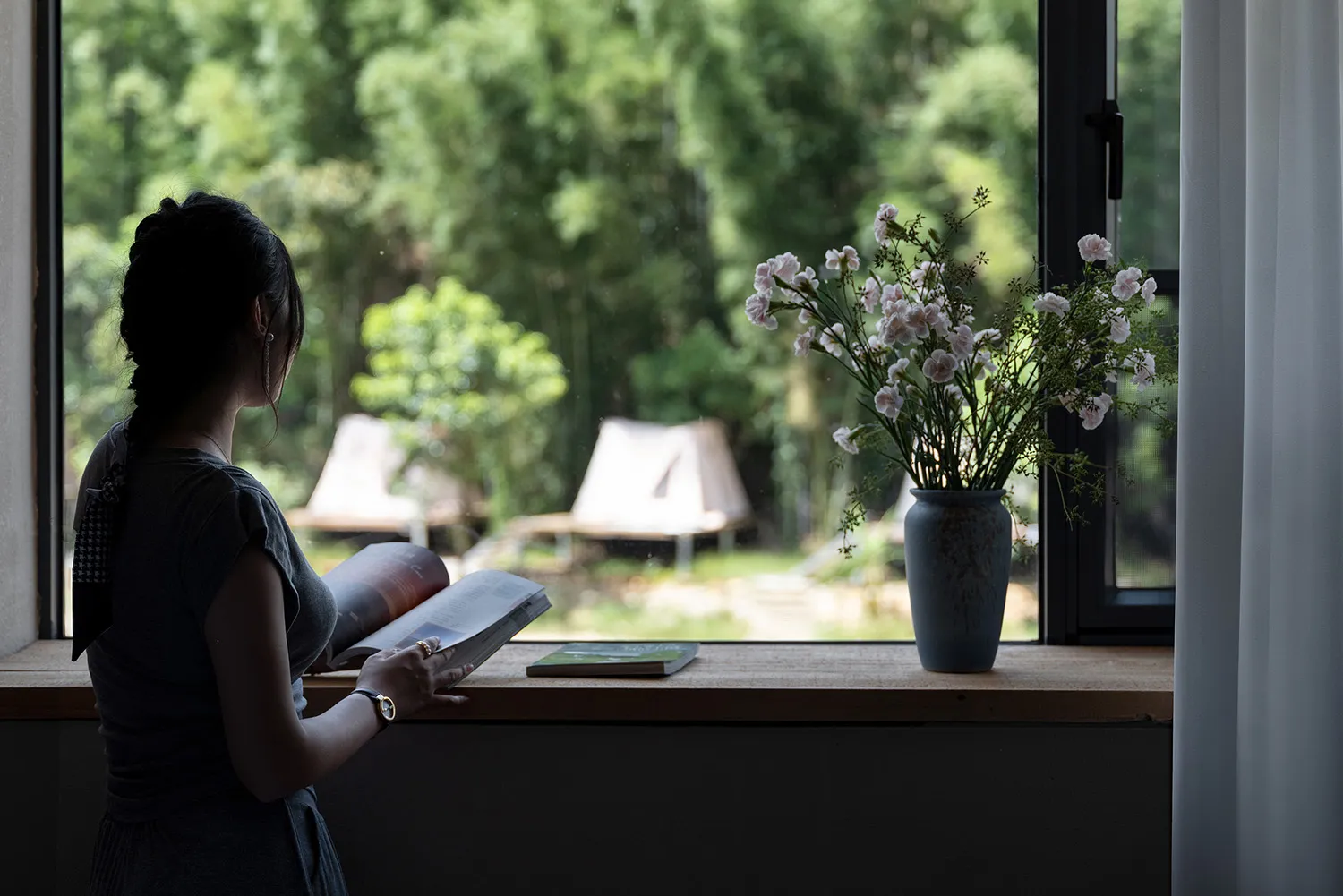 Woman reading a book by a large window overlooking a green forest, with a vase of pink flowers on the wooden windowsill.