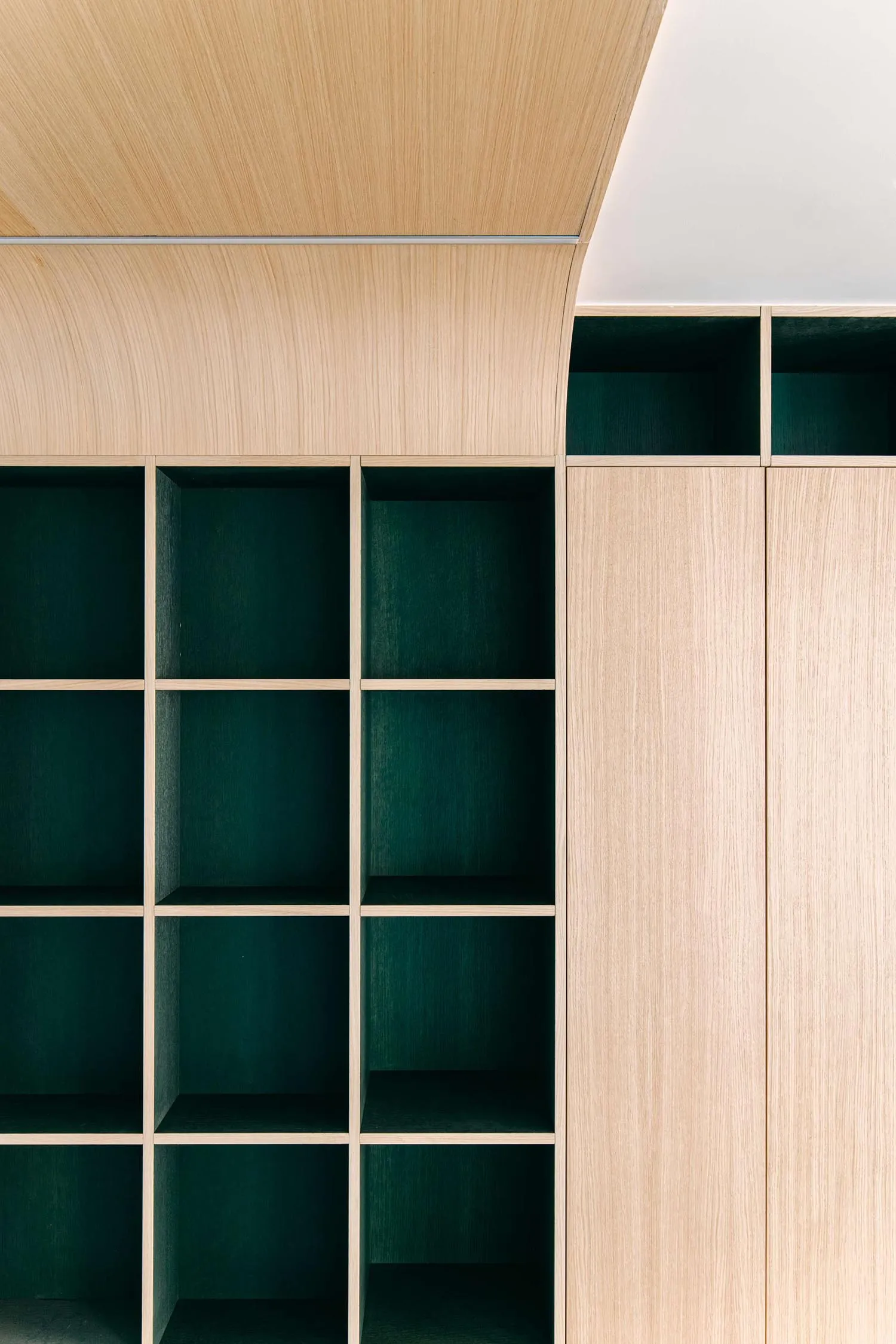Close-up of a modern built-in bookshelf made of light ash wood with deep emerald green painted interior compartments and a curved upper section.