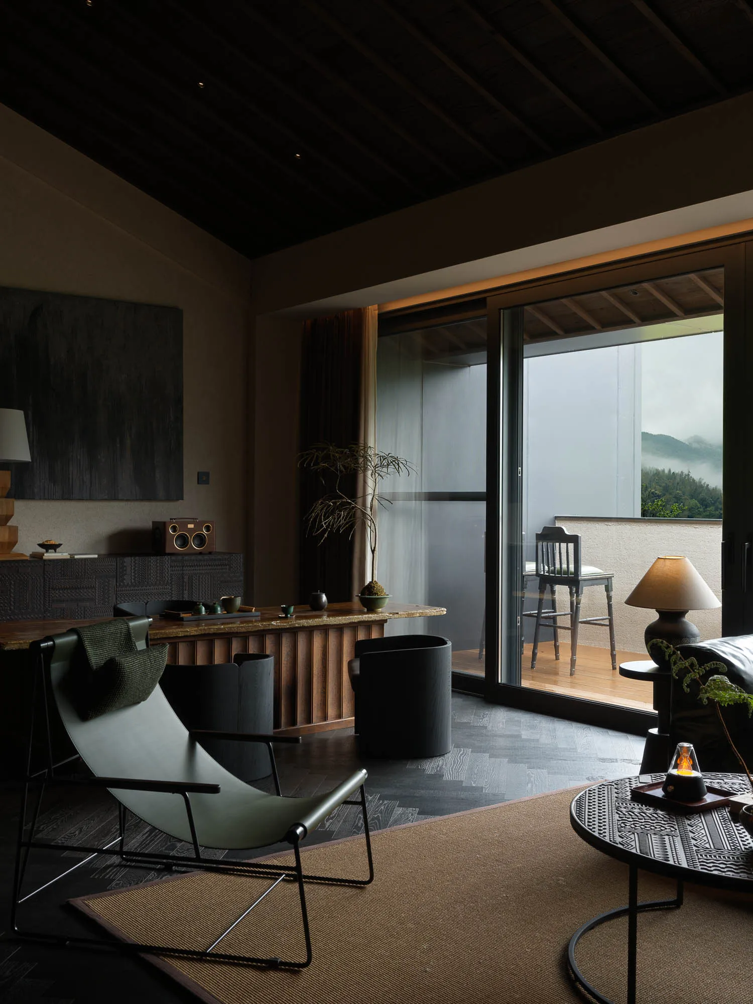Modern living room featuring a dark wood ceiling, olive green sling chair, wooden console, and large glass doors opening to a balcony with mountain views.
