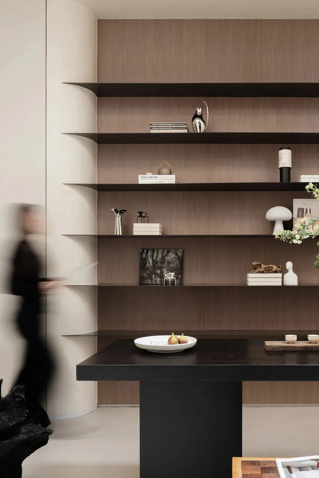 Modern living room with dark walnut floating shelves displaying books, sculptures, and decorative objects against a light neutral wall, with a black console table in the foreground.