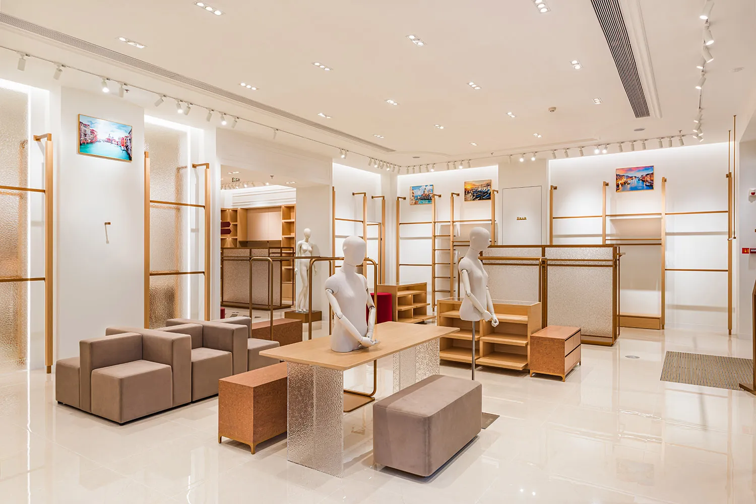 現代簡約時裝精品店室內，設有米色模組沙發、淺橡木展示桌搭配紋理玻璃基座、白色人型模特兒、黃銅層架，以及光亮的白色磁磚地板。