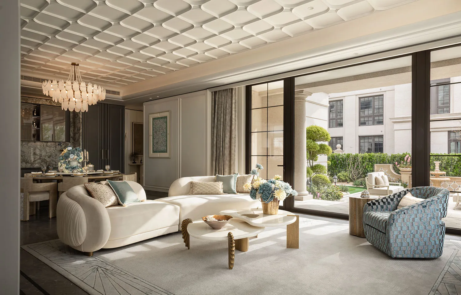 Bright and airy living room featuring a curved cream sofa, blue patterned armchair, organic-shaped marble coffee table, and large glass doors opening to a landscaped garden.