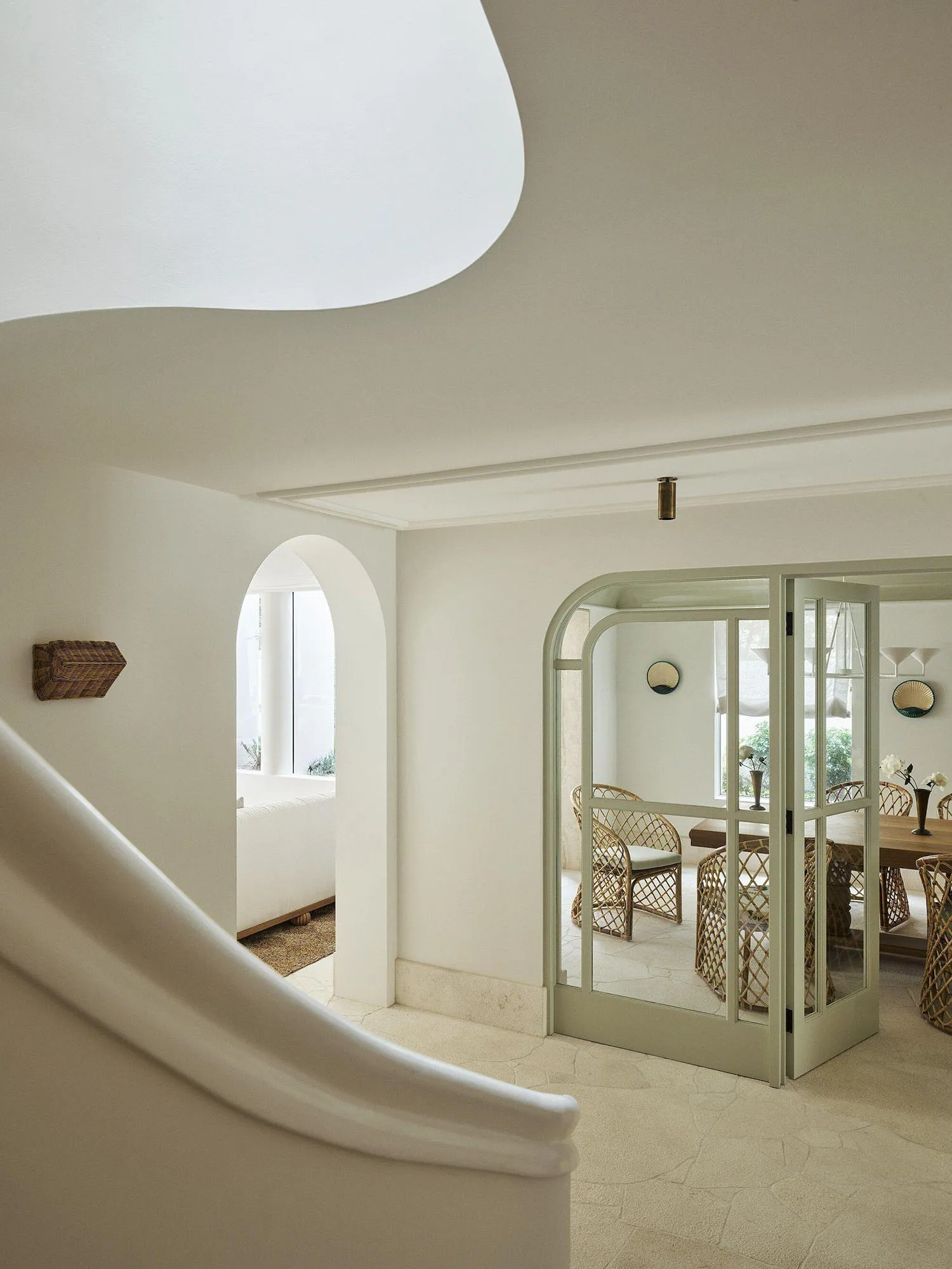 View from a curved staircase into a bright dining room with sage green arched French doors, rattan dining chairs, and white plaster walls.