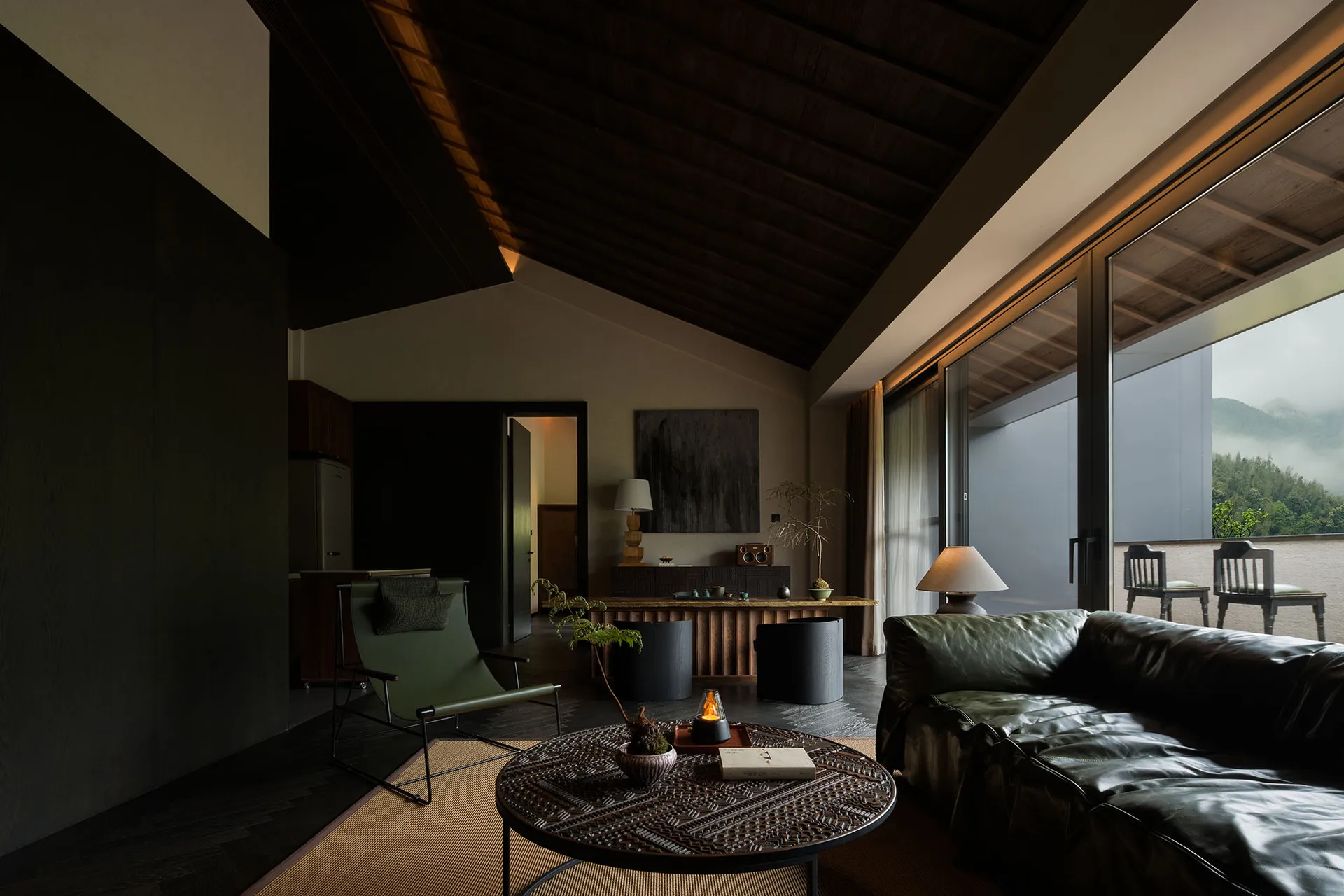 Modern living room featuring a dark wood ceiling and walls, a dark green leather sofa, a matching green lounge chair, an intricately carved round coffee table, and large windows overlooking misty mountains.