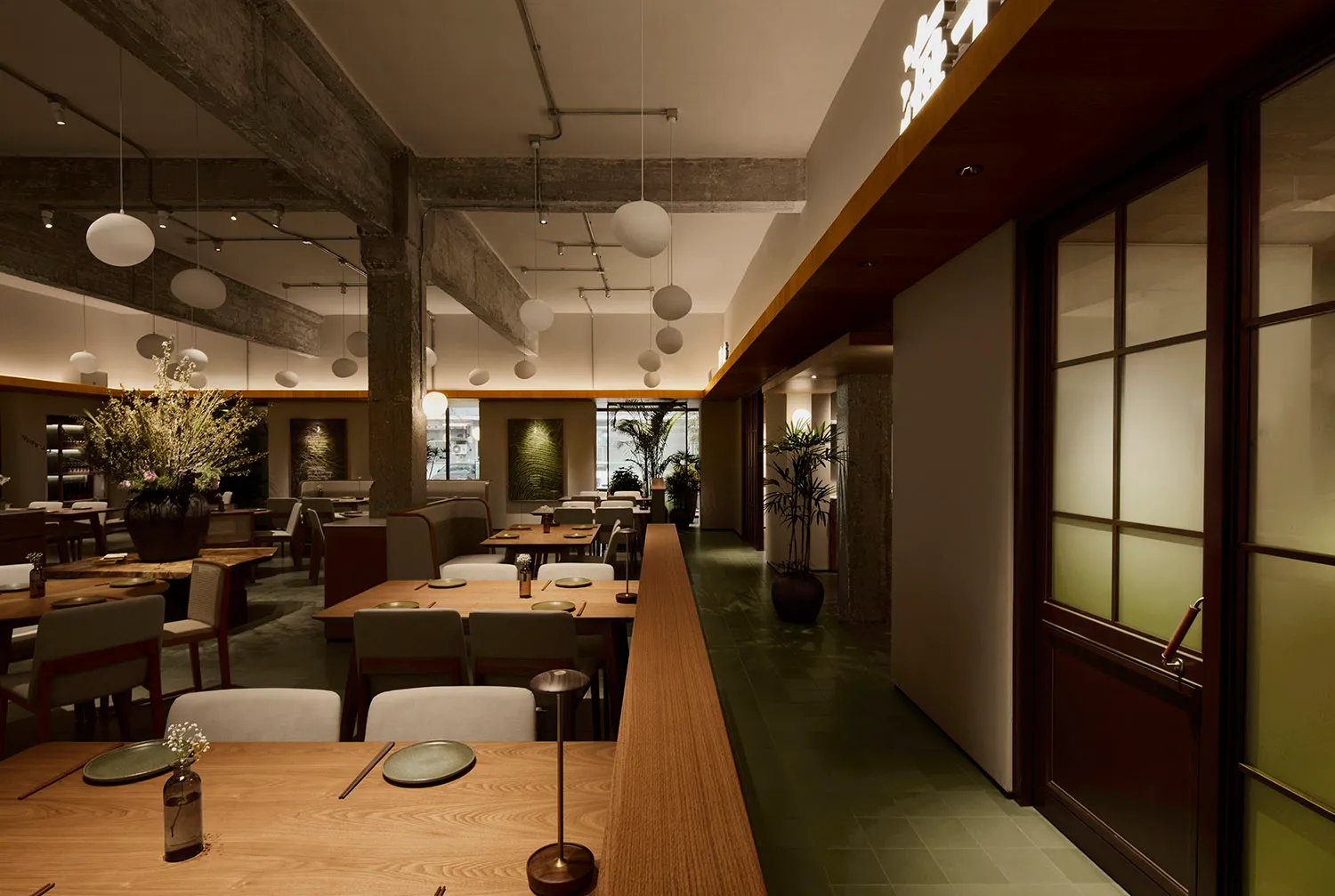 Interior view of a modern Japanese restaurant featuring long light oak dining tables, upholstered chairs, exposed concrete beams, and hanging spherical pendant lights.