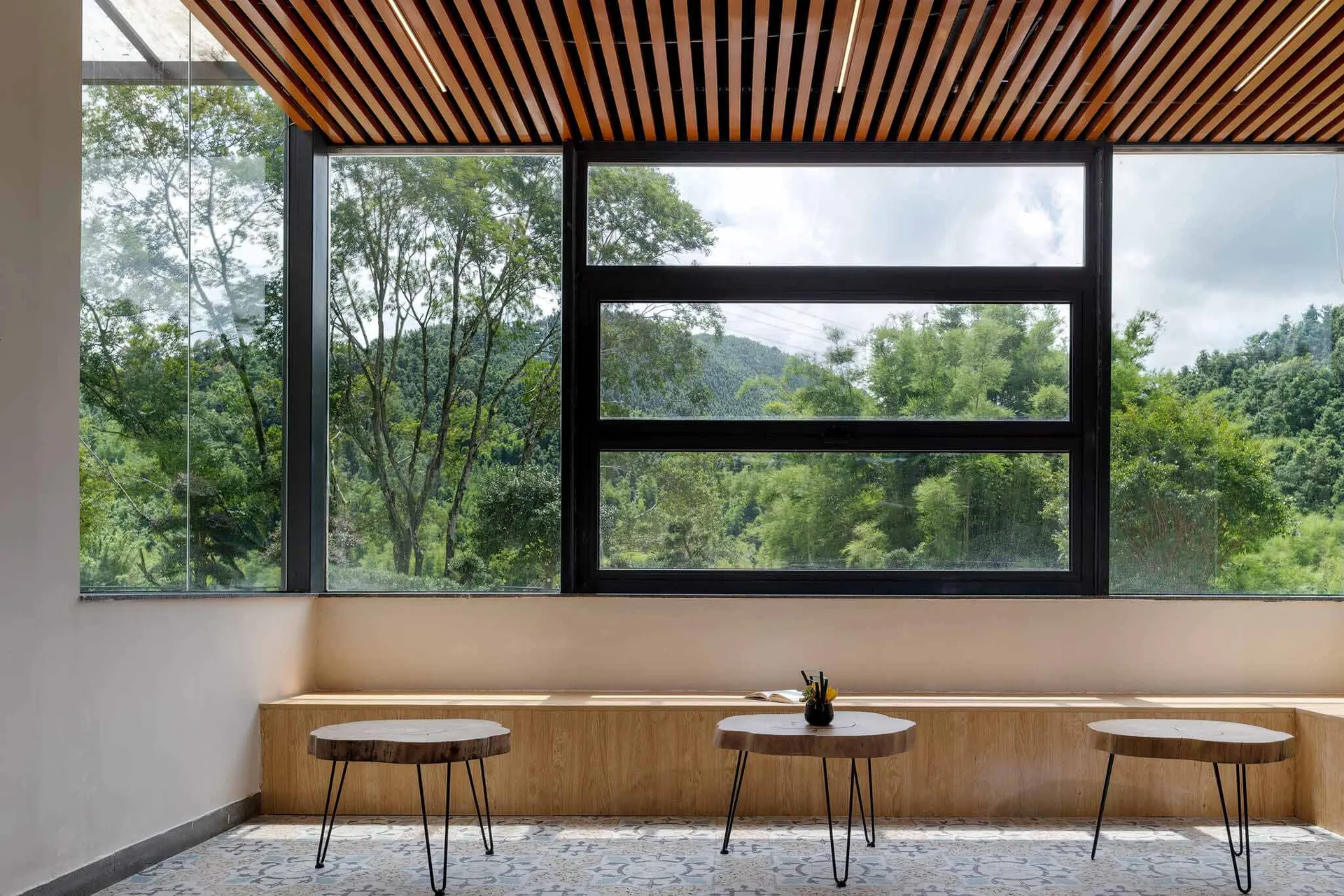 Modern sunroom interior with live-edge wood slab tables on hairpin legs, built-in window bench, patterned tile floor, and expansive black-framed windows revealing a dense forest and mountain landscape.