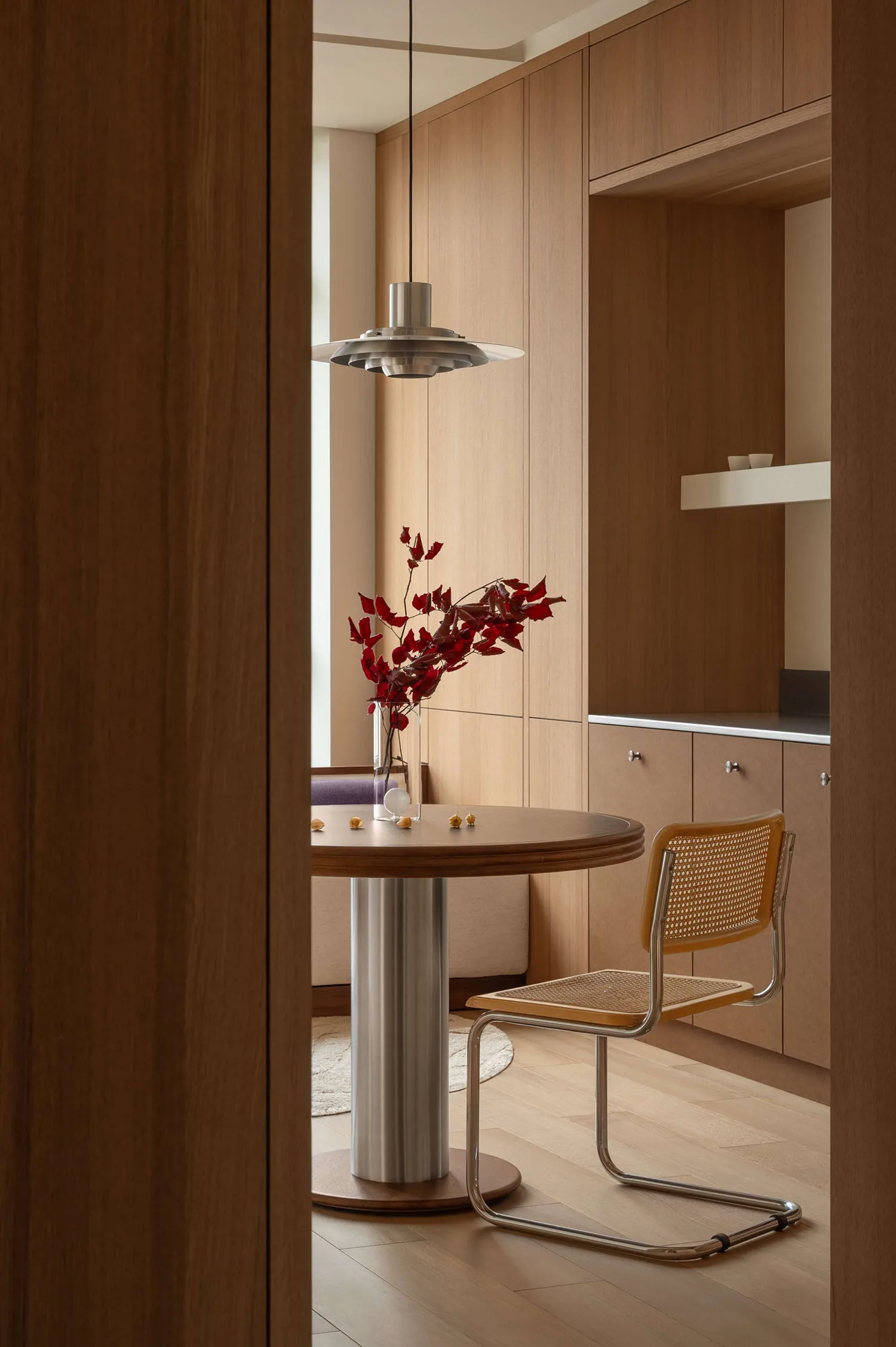 Modern minimalist breakfast nook featuring light walnut cabinetry, a round pedestal dining table, a cane and chrome chair, and a Louis Poulsen PH 5 pendant lamp with red floral arrangement.