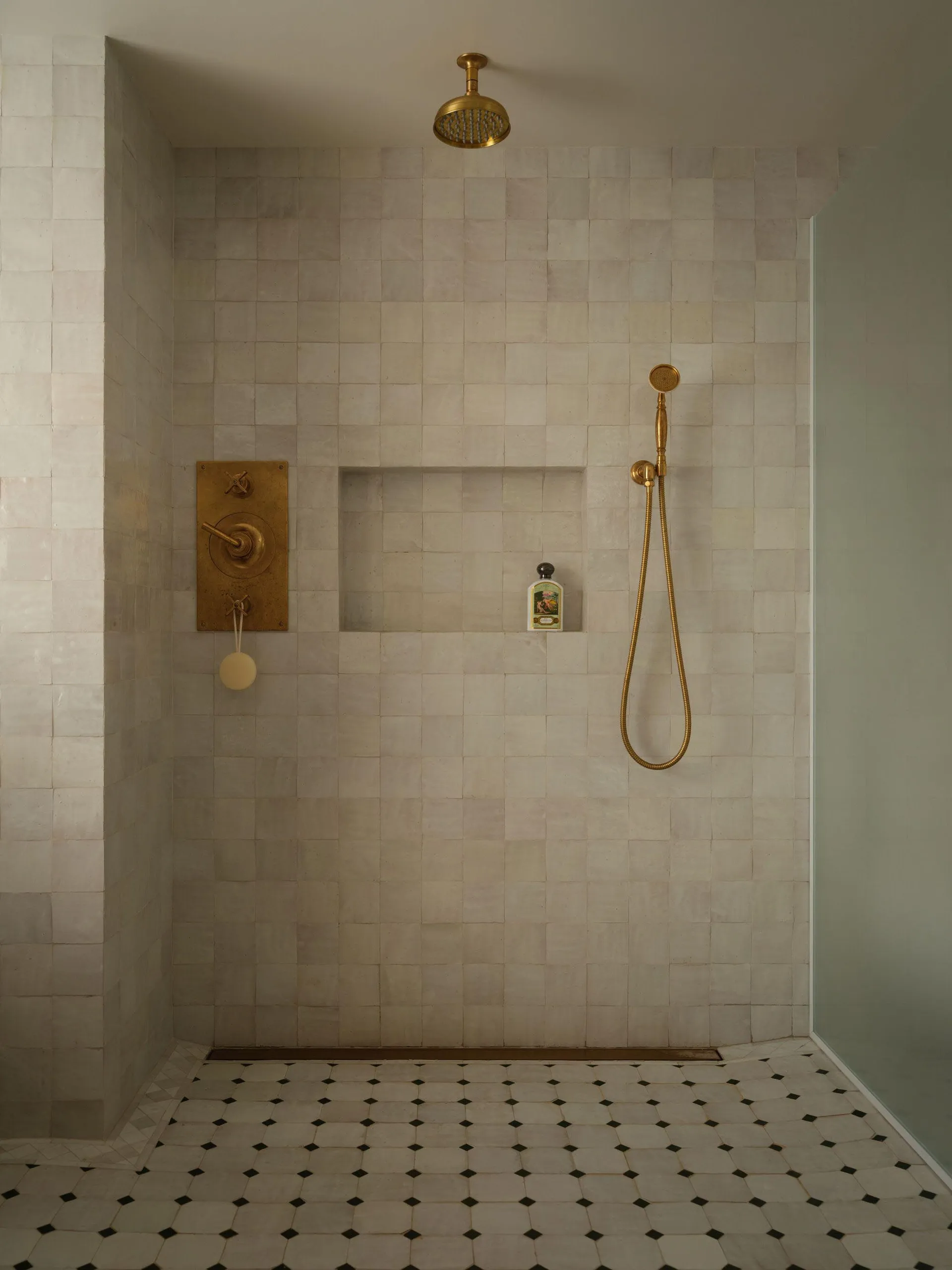 Minimalist shower space with off-white zellige tiled walls, a recessed niche holding a bath product, unlacquered brass shower head and mixer, and geometric black-and-white mosaic floor tiles.