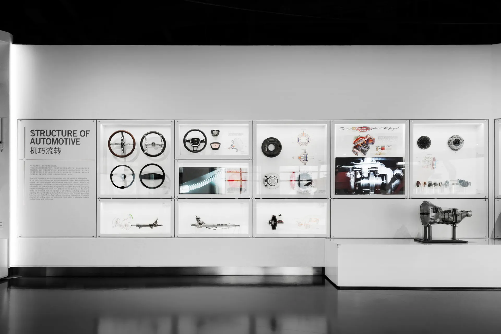 Museum exhibition wall displaying various automotive parts including steering wheels, driveshafts, brakes, and engine components in illuminated acrylic cases with technical diagrams and explanatory text.