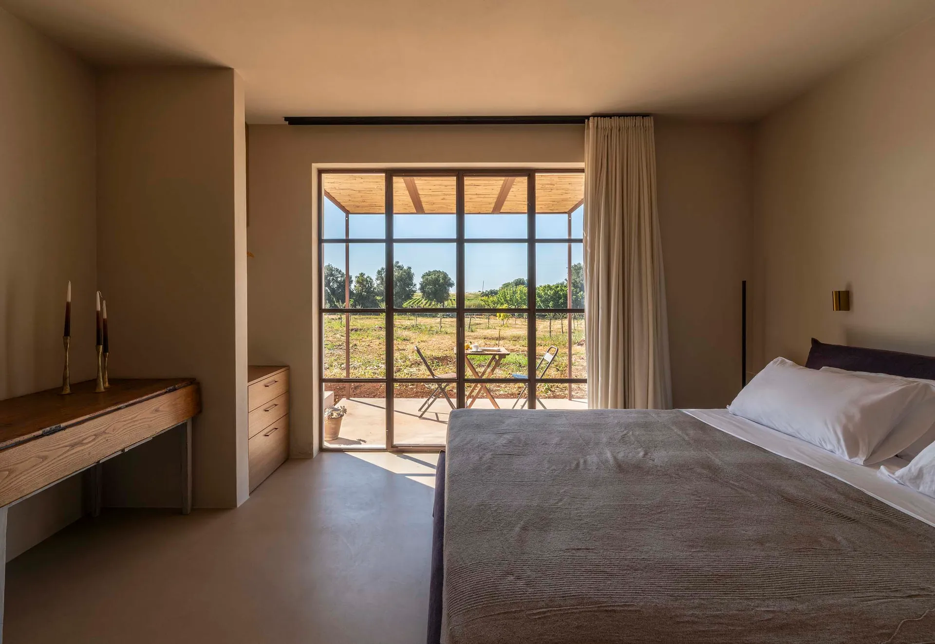 Modern minimalist bedroom featuring a large bed with grey bedding, wooden console desk, and floor-to-ceiling glass doors opening to a vineyard terrace.