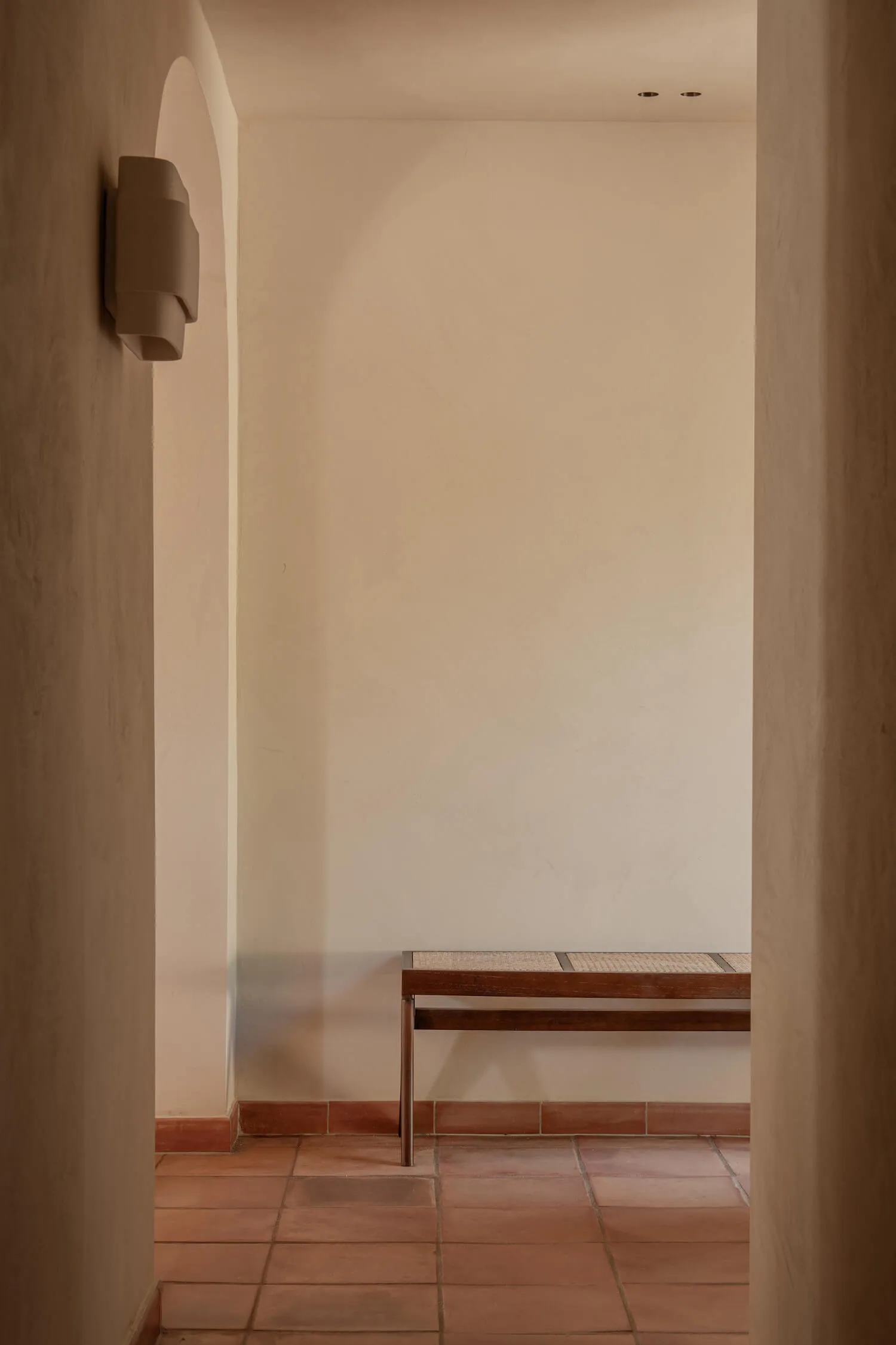 Minimalist entryway featuring terracotta floor tiles, a wooden bench with woven seat, and a sculptural wall sconce under an arched alcove.
