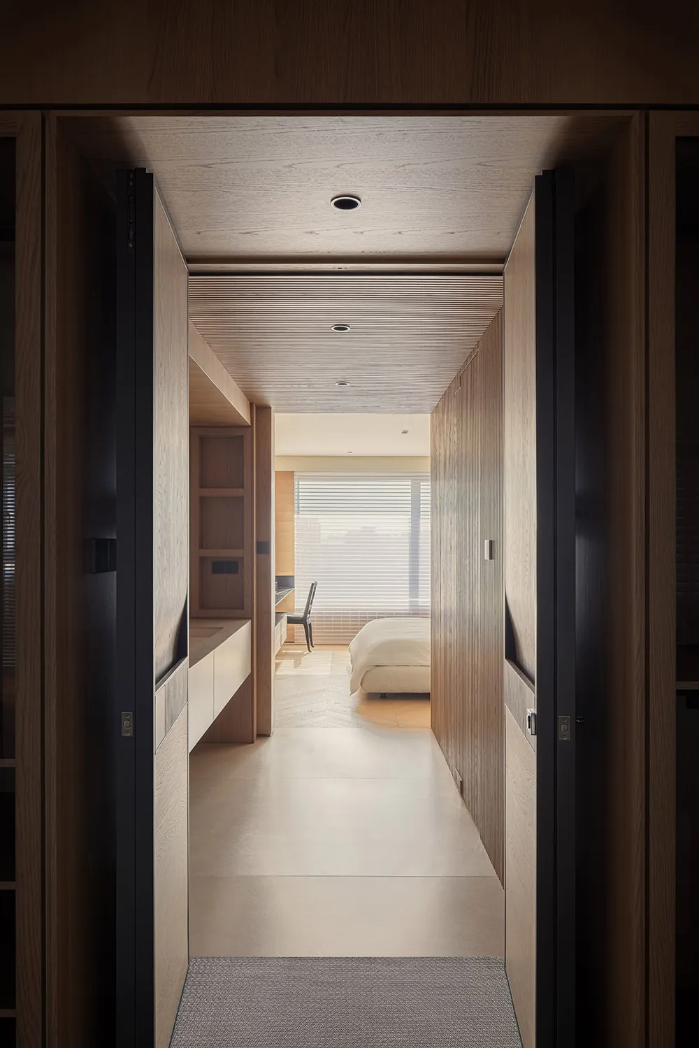 View from a doorway looking down a minimalist hallway with oak wood walls, built-in storage, and a wood slat ceiling leading to a bright bedroom with a large window.