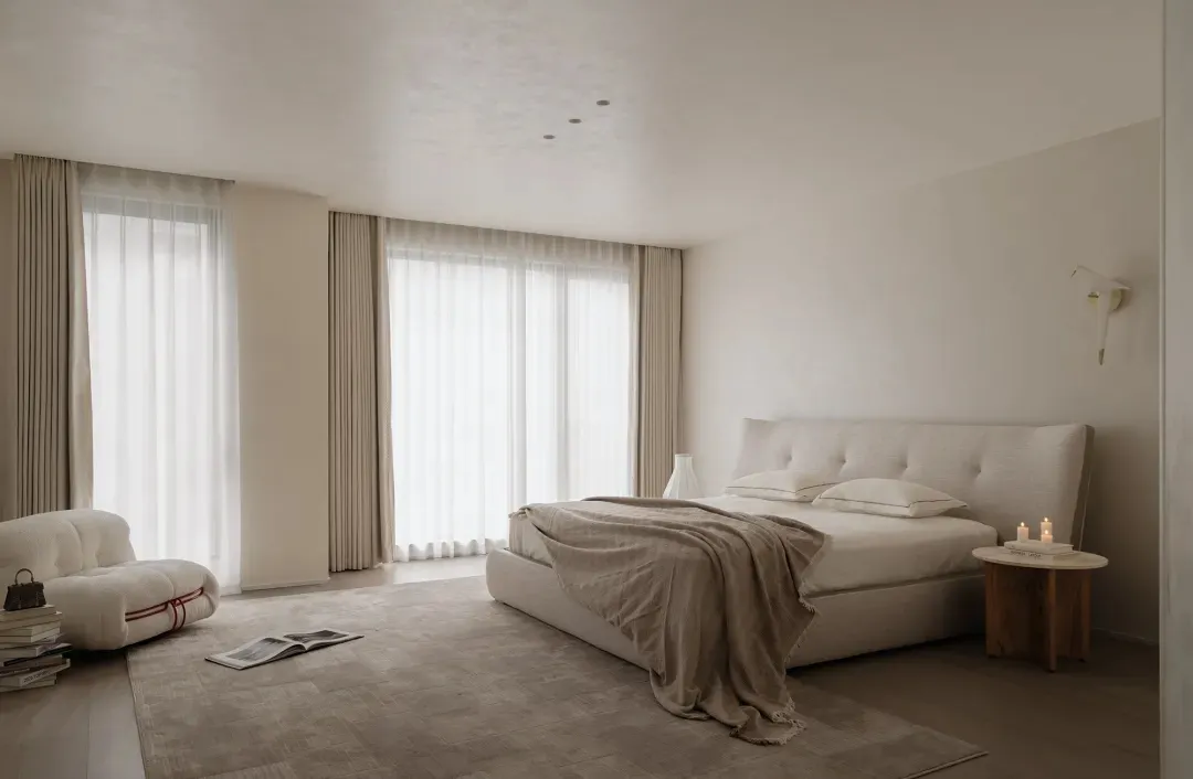 Minimalist bedroom with a large upholstered bed dressed in beige linen, a curved white bouclé sofa, sheer curtains allowing soft daylight, and a wooden side table with candles.