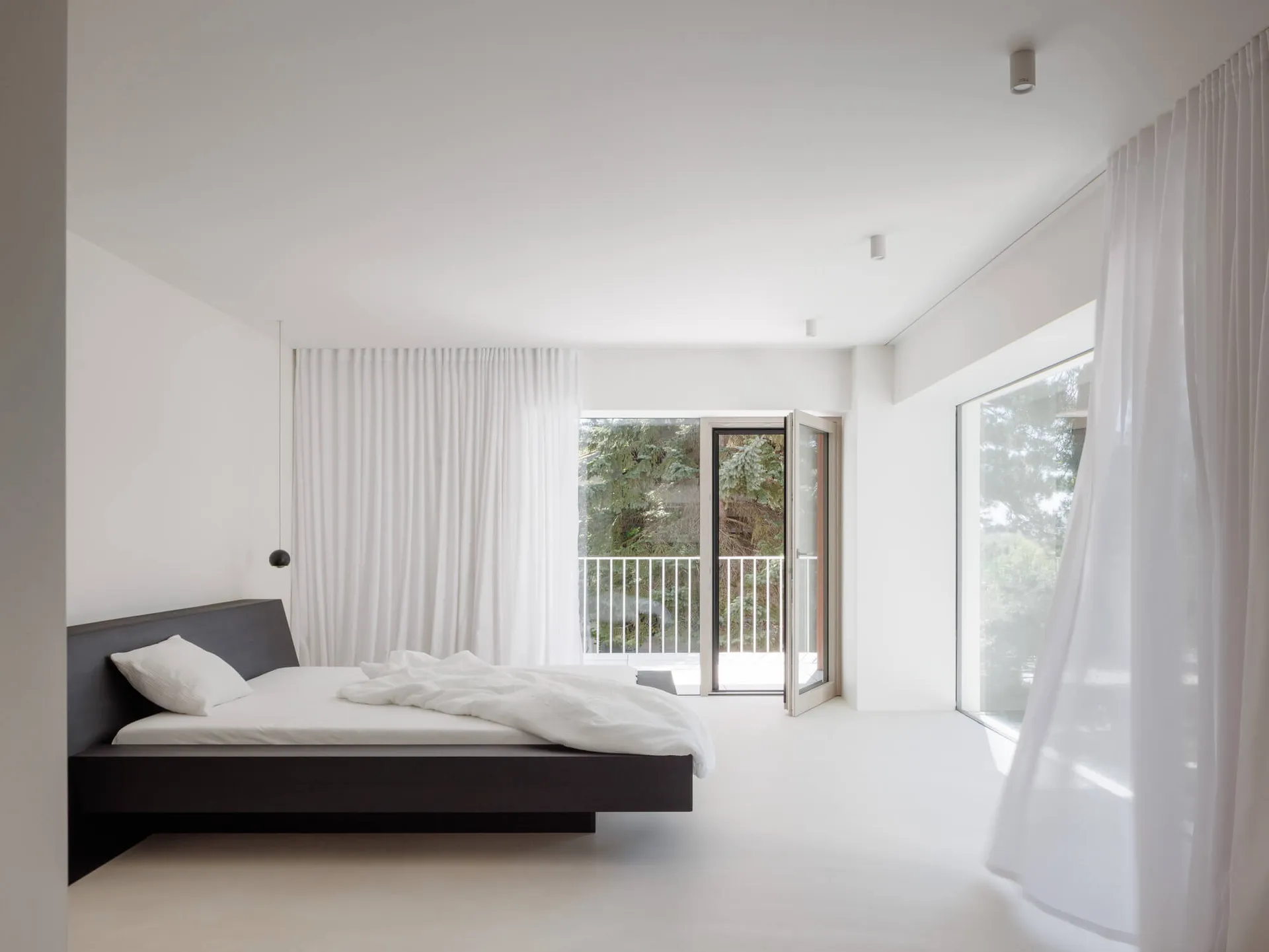 Modern minimalist bedroom with a dark wood floating platform bed, white bedding, sheer white curtains, and large glass doors opening to a balcony with greenery.
