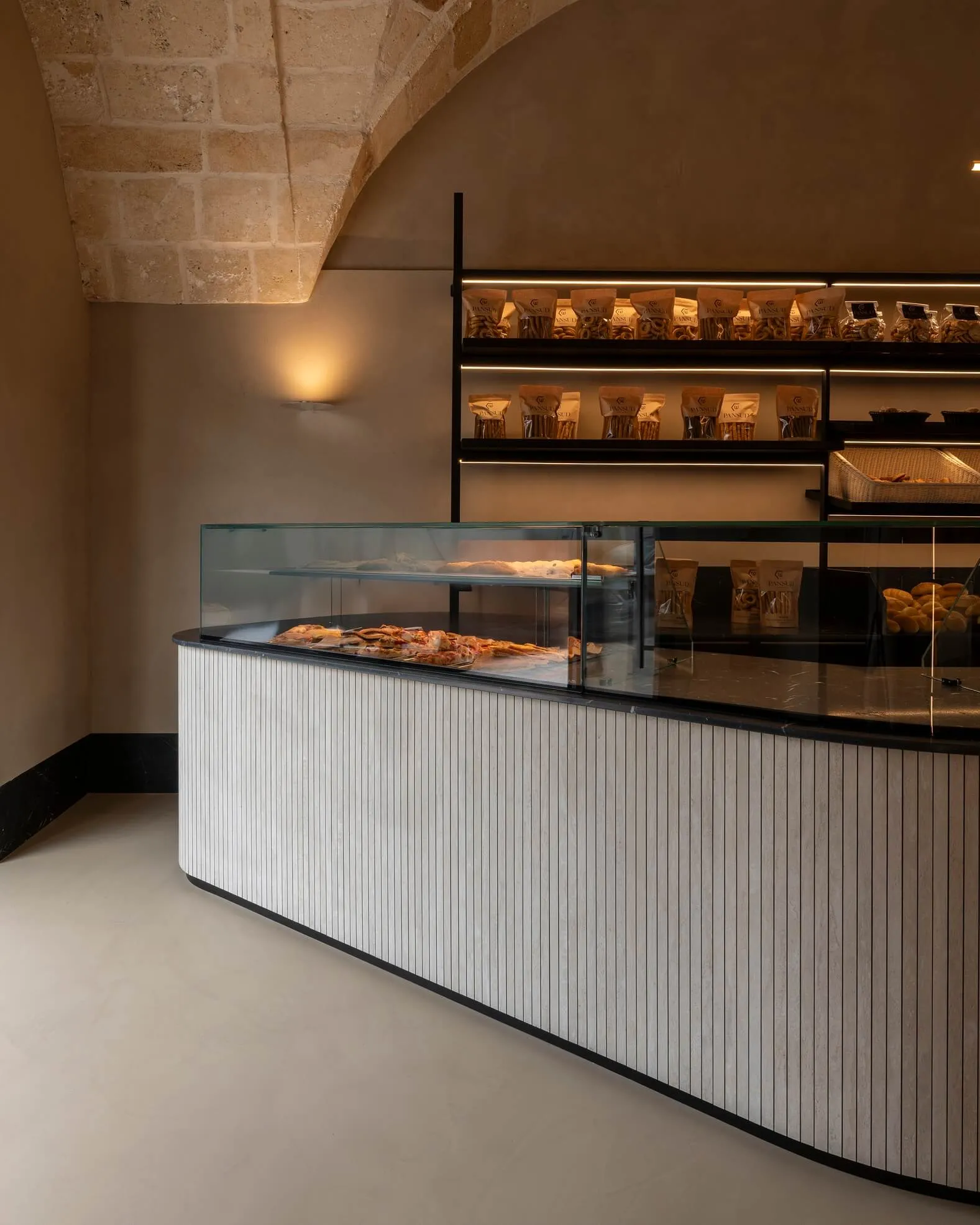 Modern bakery interior featuring a curved white fluted counter with glass display case, black marble top, and black metal shelving filled with packaged goods against a stone arched ceiling.