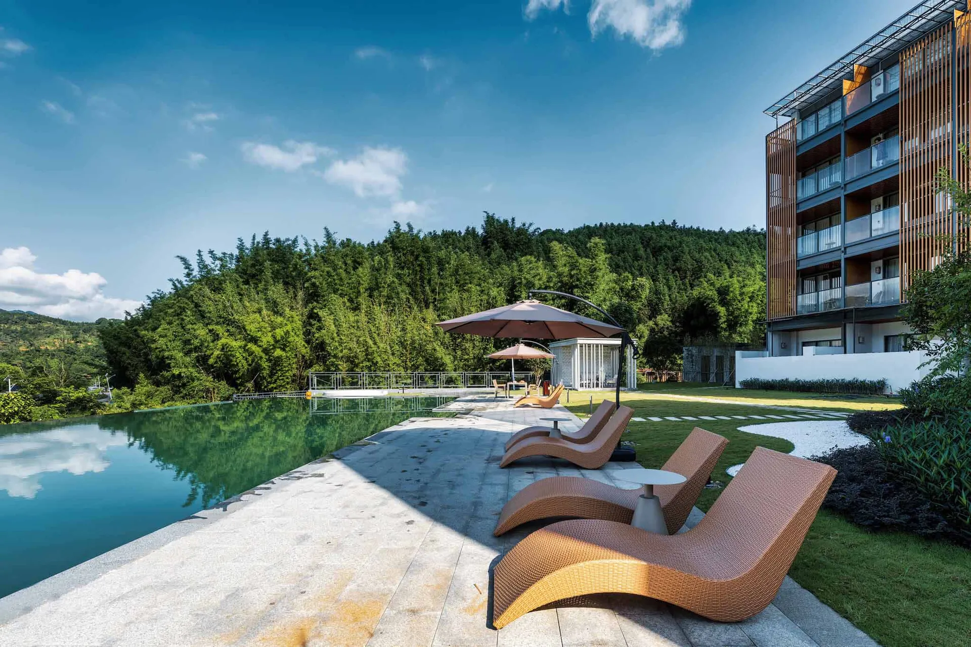 Modern woven rattan lounge chairs arranged on a stone terrace beside a reflecting pool, with a contemporary resort building and dense green bamboo forest in the background under a clear blue sky.